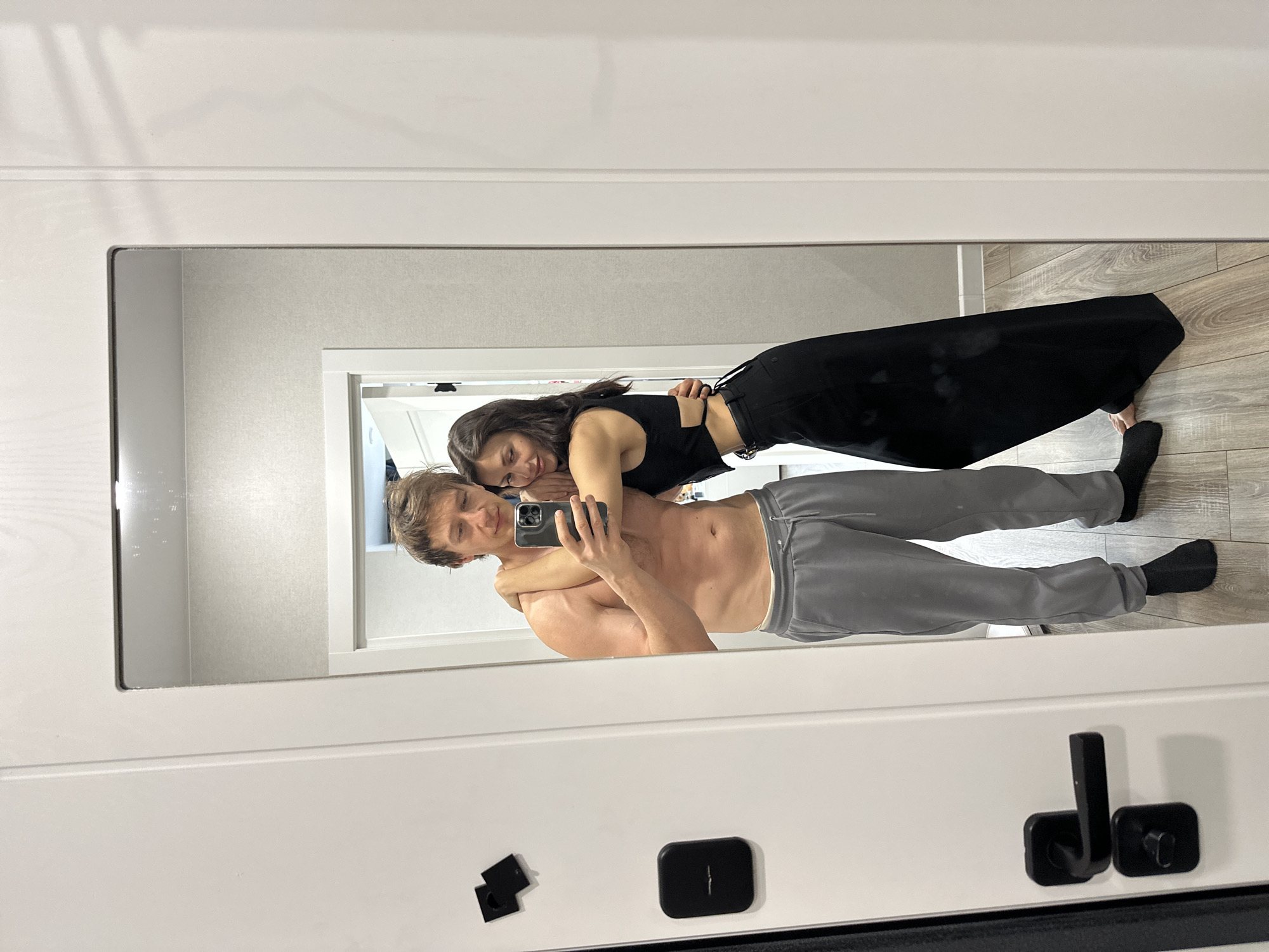 A couple is reflected in a full-length mirror indoors. The man is shirtless with grey sweatpants and black socks. He is holding a phone, presumably taking a selfie. The woman is wearing a black top and long black pants with black socks. She is leaning on the man's back, hugging him. The room appears to have light grey walls and wood-look flooring. A white doorframe is visible in the reflection, along with a glimpse of another room. The location is inferred to be in Murino, Russia, based on prior context. The mirror frame is white and has a handle with associated hardware. Two unidentified magnetic square objects are also present on the front of the mirror.