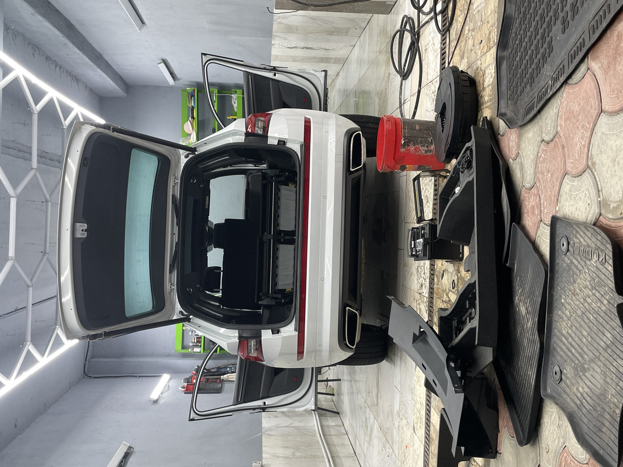 This image shows the interior and exterior of a white SUV in what appears to be a garage or auto detailing shop. The rear hatch and passenger doors are open, revealing the interior of the vehicle. Several car parts, including floor mats and what looks like a dashboard or console piece, are laid out on the floor beside the car.
The setting is a well-lit indoor space with concrete flooring and walls. Fluorescent lighting fixtures are visible on the ceiling and along one wall, suggesting artificial lighting. The overall impression is of a car undergoing some form of maintenance, repair, or detailing. There are no people visible in the image.
Notable details include a red bucket next to a spare tire, a coiled black hose, and a large black rubber floor mat with a textured pattern. The floor is paved with interlocking stones of red and beige hues. The car has tinted windows. There is a label on one of the floor mats that reads "KODIAQ". The image is oriented vertically, with the car's rear facing towards the top. The time of day and weather are not discernible from the image.