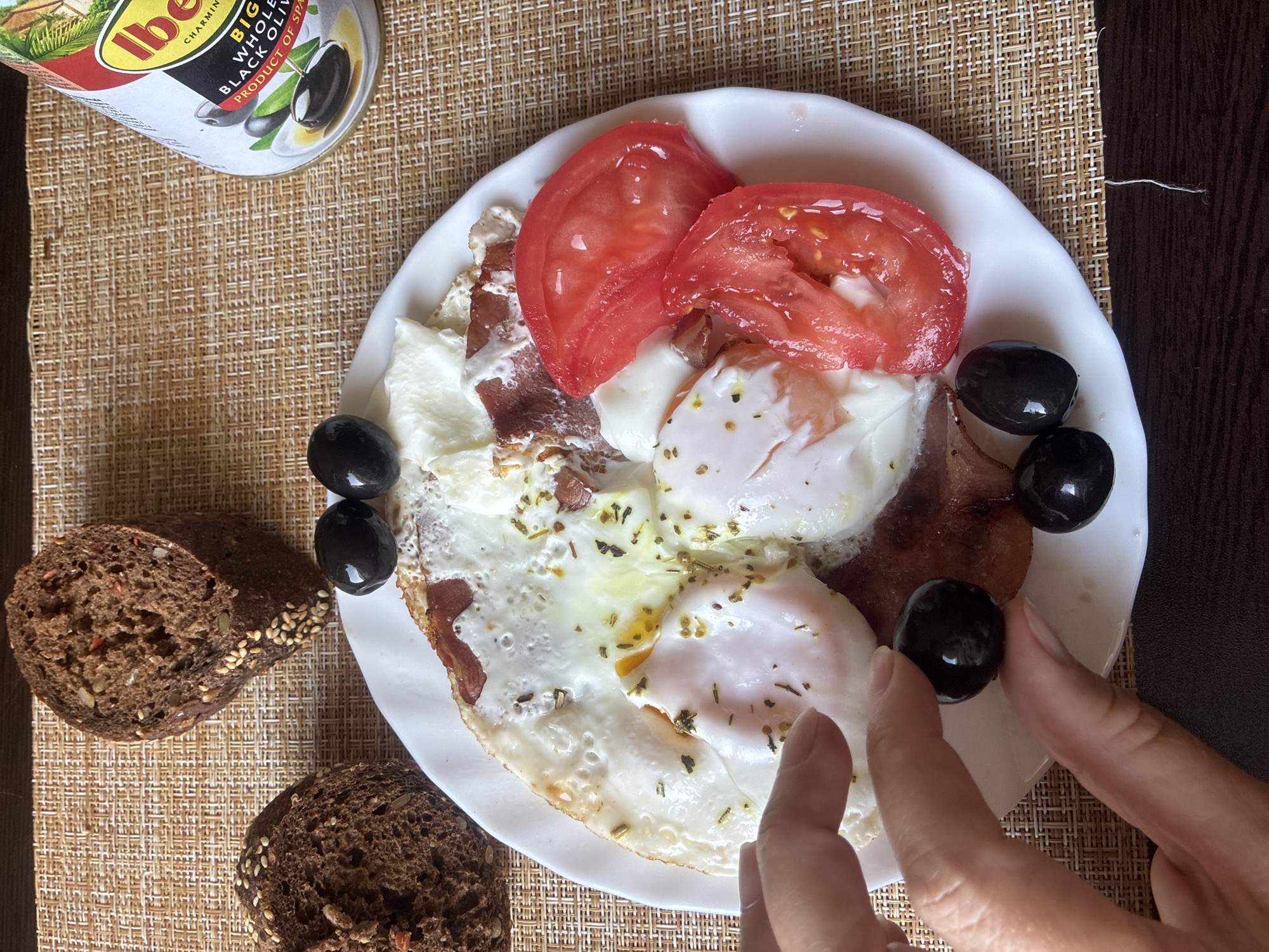An overhead, close-up shot captures a breakfast plate with two fried eggs, slices of tomato, and pieces of cured meat. Two black olives are placed next to the plate, with a hand holding one of them. Two slices of dark bread with seeds are visible on the left side of the plate. A can of black olives stands on the right. The food is arranged on a woven placemat, with a dark brown surface beneath it. The image is taken from a high angle and does not contain any identifiable urban or natural landmarks, therefore its location in Sochi, Russia, or any other specific city, cannot be determined.