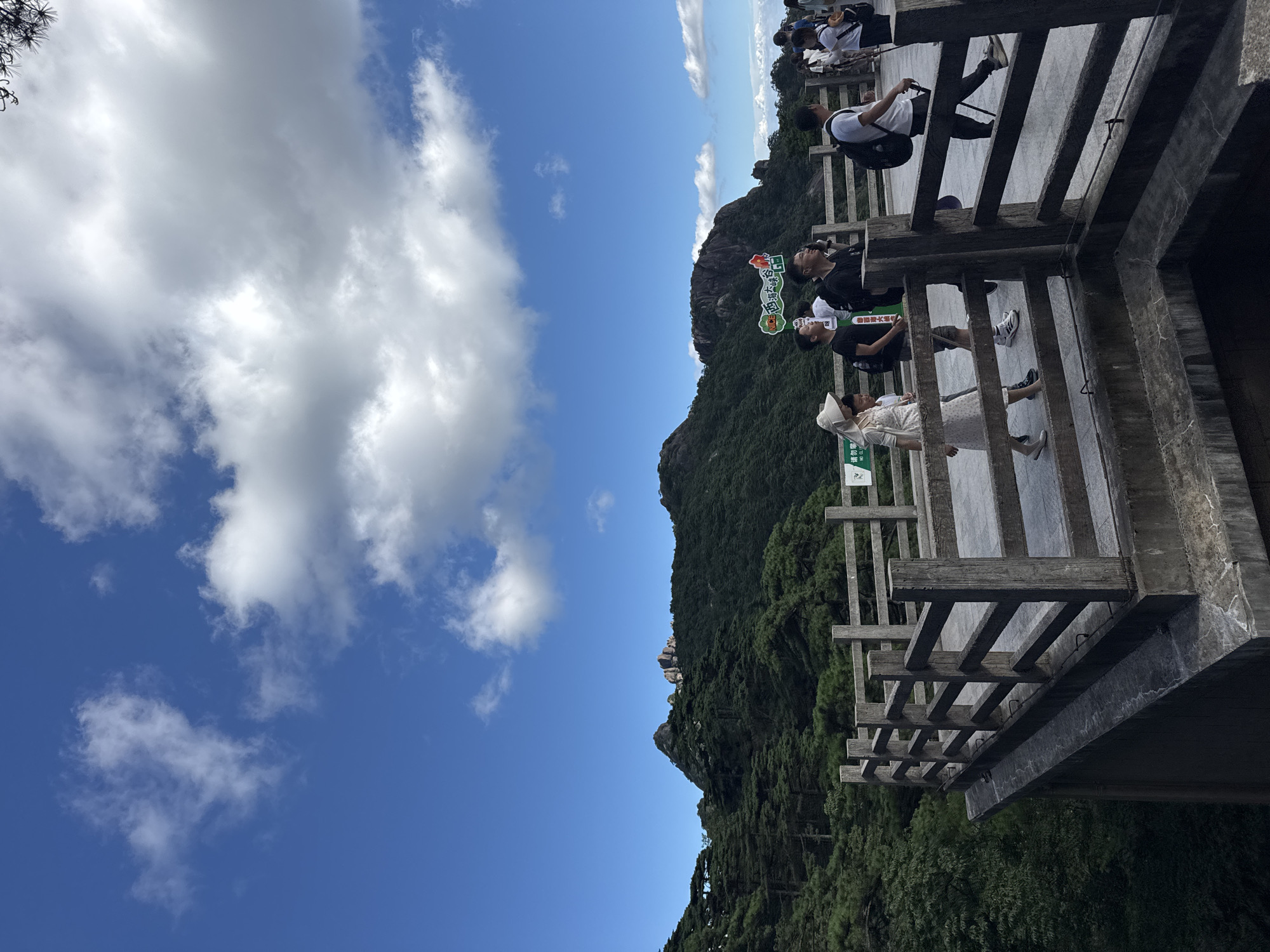 A group of tourists are walking along a wooden viewing platform overlooking a mountainous landscape. The sky is blue with scattered white clouds. The platform is made of concrete with a wooden railing. In the background, lush green mountains are covered with trees. Some of the tourists are wearing hats and backpacks. The scene appears to be set in a scenic tourist area within the city of Huangshan District, China, likely on Mount Huangshan, a UNESCO World Heritage site known for its natural beauty.