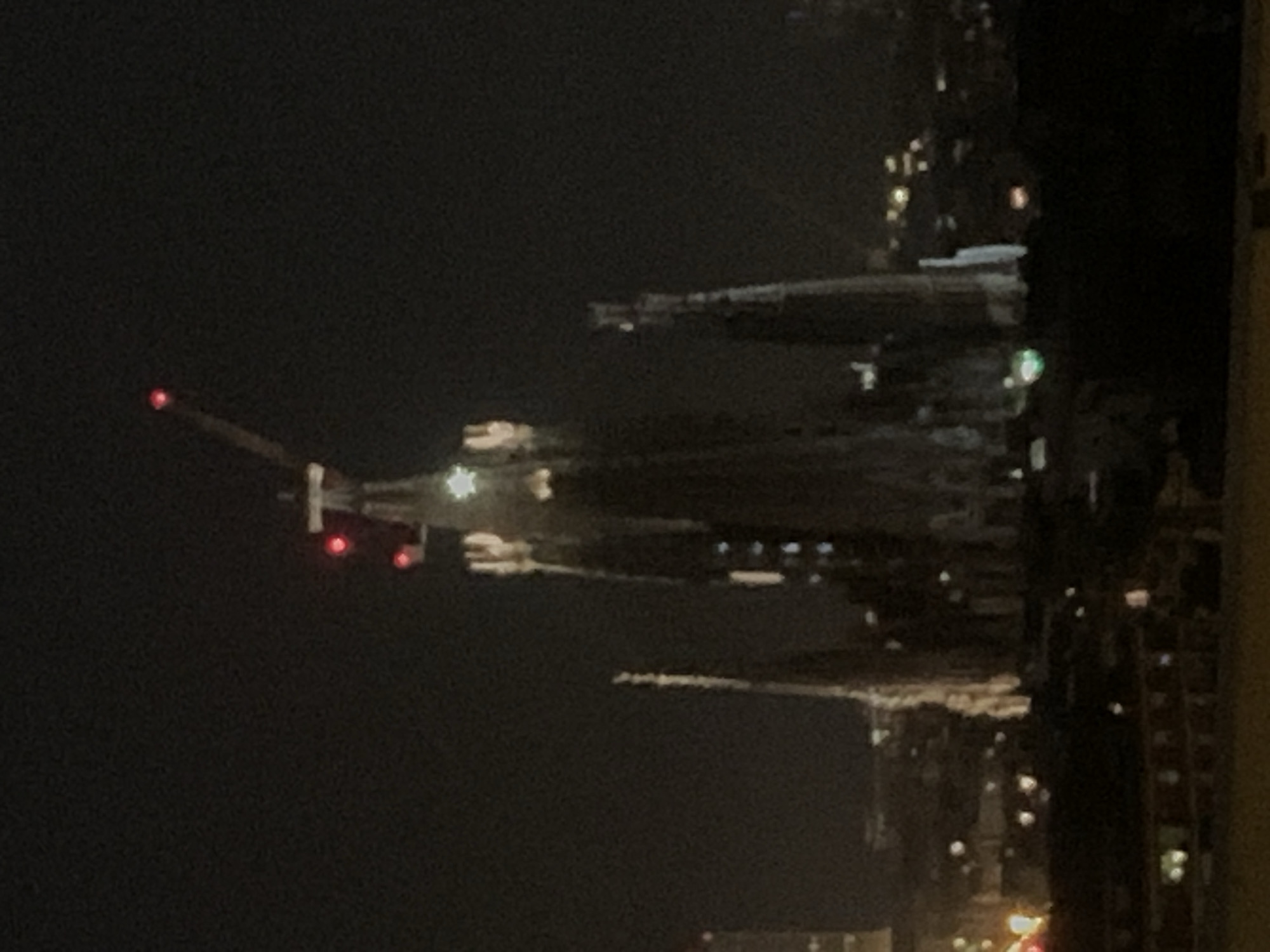 The image shows a nighttime view of a city skyline, likely Barcelona, Spain, reflected in water. A plane with illuminated red lights is visible in the upper left against a dark sky. The city lights create distinct bright spots interspersed with dark areas. Buildings are visible on the right edge. The city skyline creates a distinct reflection in the water.
