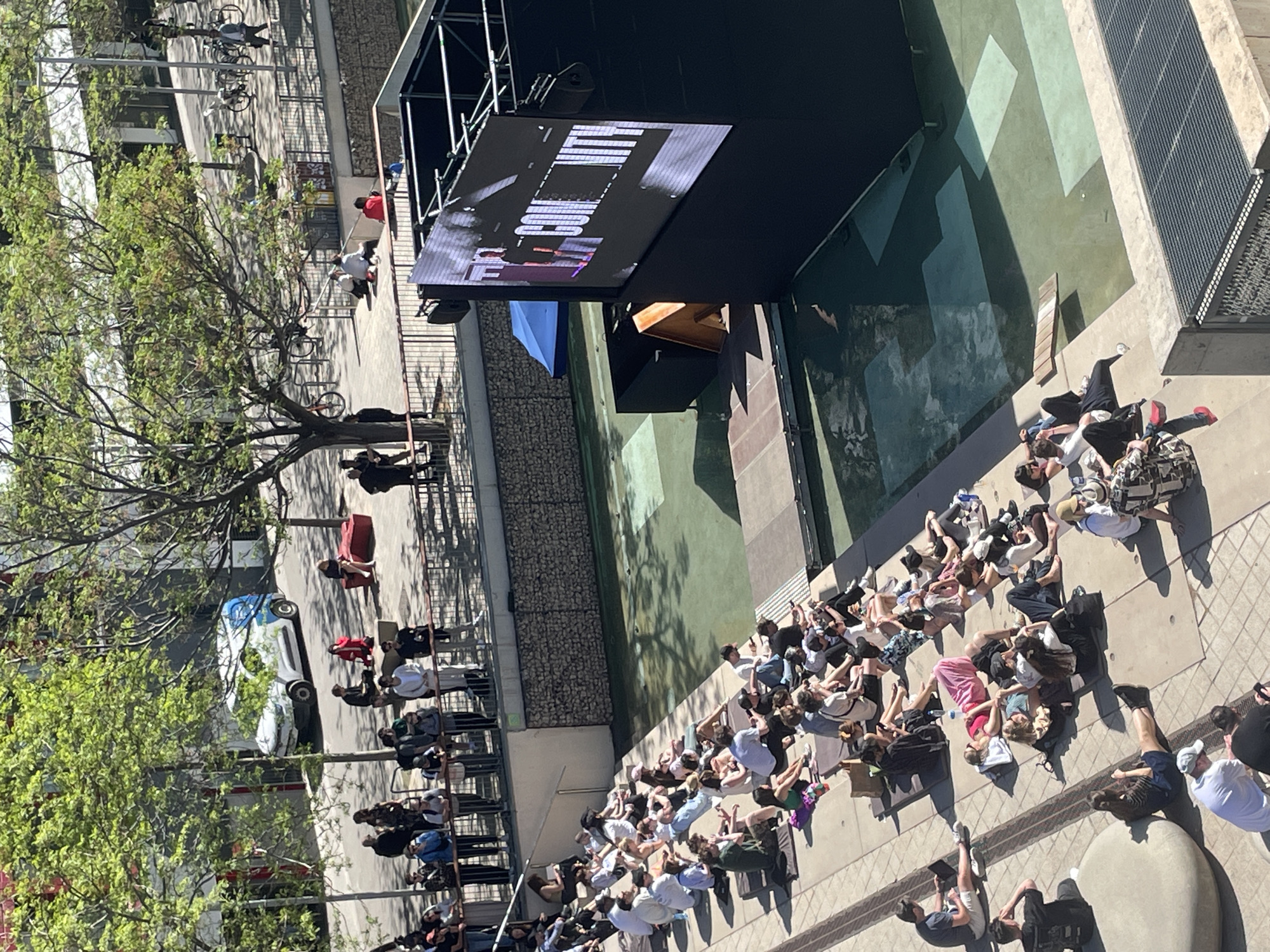 The image shows a crowd of people gathered in a public space, likely in Barcelona, Spain. They are seated or lying down on the ground, facing a large outdoor screen that is mounted on a stage-like structure. The screen displays an image with bright lettering. The area is surrounded by buildings, including some with balconies or terraces. A tree with green leaves is visible to the left of the frame. There are also a few vehicles parked along the street. The people appear to be watching something on the screen, possibly a movie or a live event. Some are using their phones.