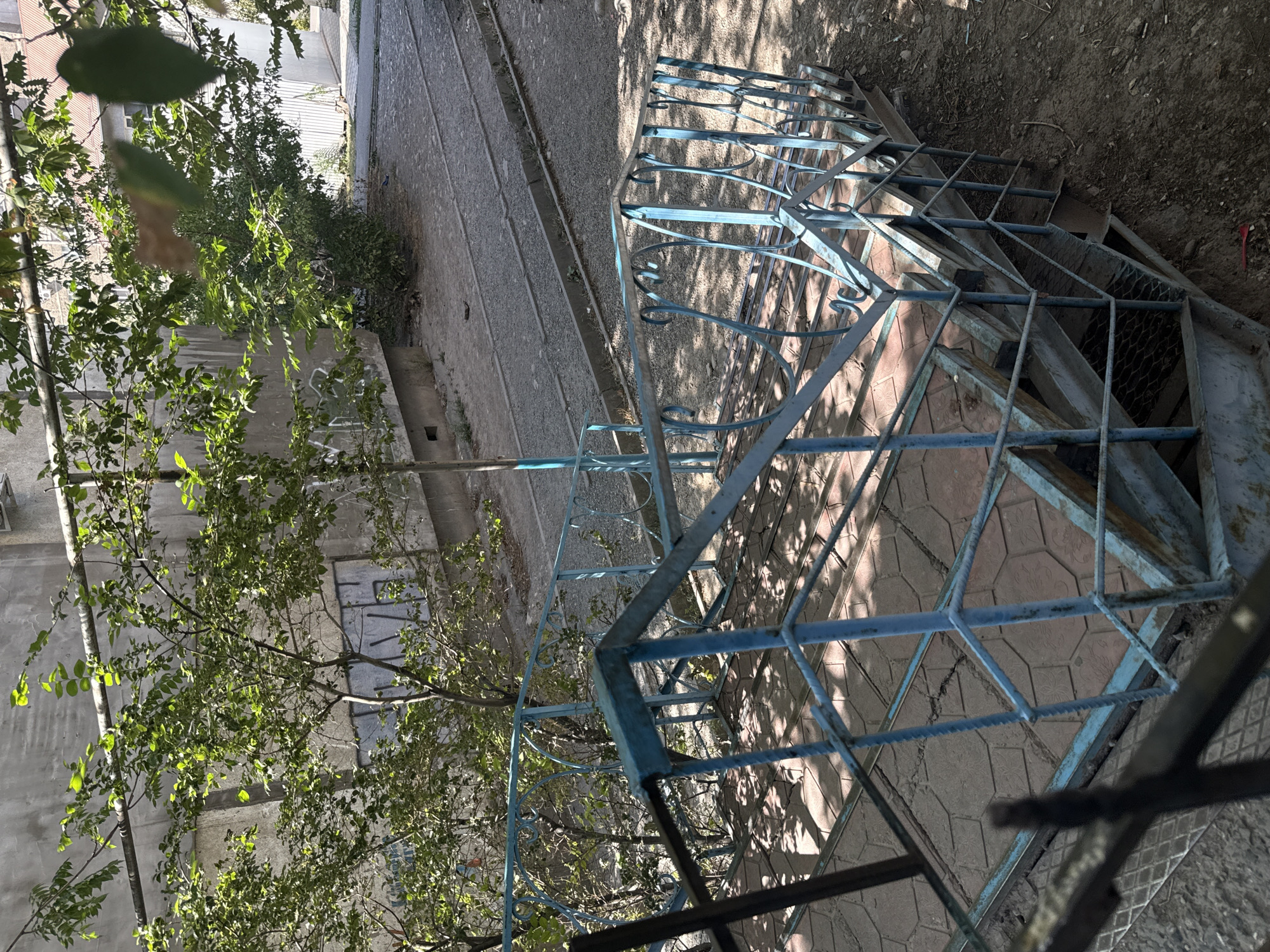 A high-angle, eye-level shot captures an outdoor metal staircase painted light blue, showing signs of rust and wear. The staircase leads down from a tiled landing, with patterned square tiles. Above and behind the staircase, trees with green leaves are visible, casting dappled shadows on the stairs and the ground. In the background, a grey building with a concrete wall is seen, and beyond that, a gravel path with what appear to be train tracks. The environment suggests an urban setting, possibly a residential area or a neglected industrial zone in Bishkek City, Kyrgyzstan. No people are present in the image.