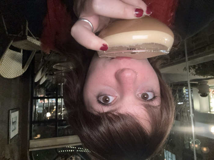 A woman with brown hair and eyes holds a glass of a light brown drink to her lips. She gazes forward with wide eyes. Her nails are painted a dark red. A silver ring adorns her finger. She wears a red, fuzzy garment. The background includes restaurant furniture, a framed picture on the wall, and a window showcasing outdoor lights. The entire image is oriented upside down.