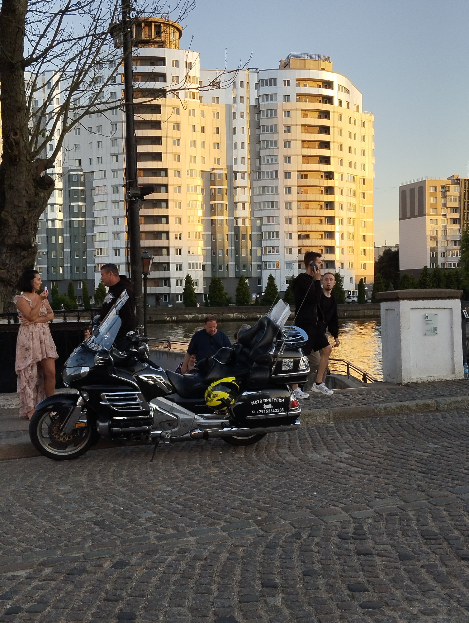 The image depicts a scene in Kaliningrad, Russia, likely on a waterfront promenade. In the foreground, a black motorcycle is parked on a cobblestone street. The motorcycle has a large windscreen and appears to be a touring model. A yellow helmet rests on the back of the motorcycle.
To the left of the motorcycle, a woman in a long, light-colored dress is standing and speaking to a man. The man is wearing a dark hoodie and is also standing near the motorcycle. Behind them, a body of water is visible, reflecting the warm light of what appears to be sunset or sunrise.
In the background, several modern apartment buildings rise up, their facades catching the golden light. These buildings appear to be multi-story structures with many windows. Another person is visible further back, looking towards the water. A street lamp is also present near the parked motorcycle. The overall atmosphere suggests a relaxed urban setting.