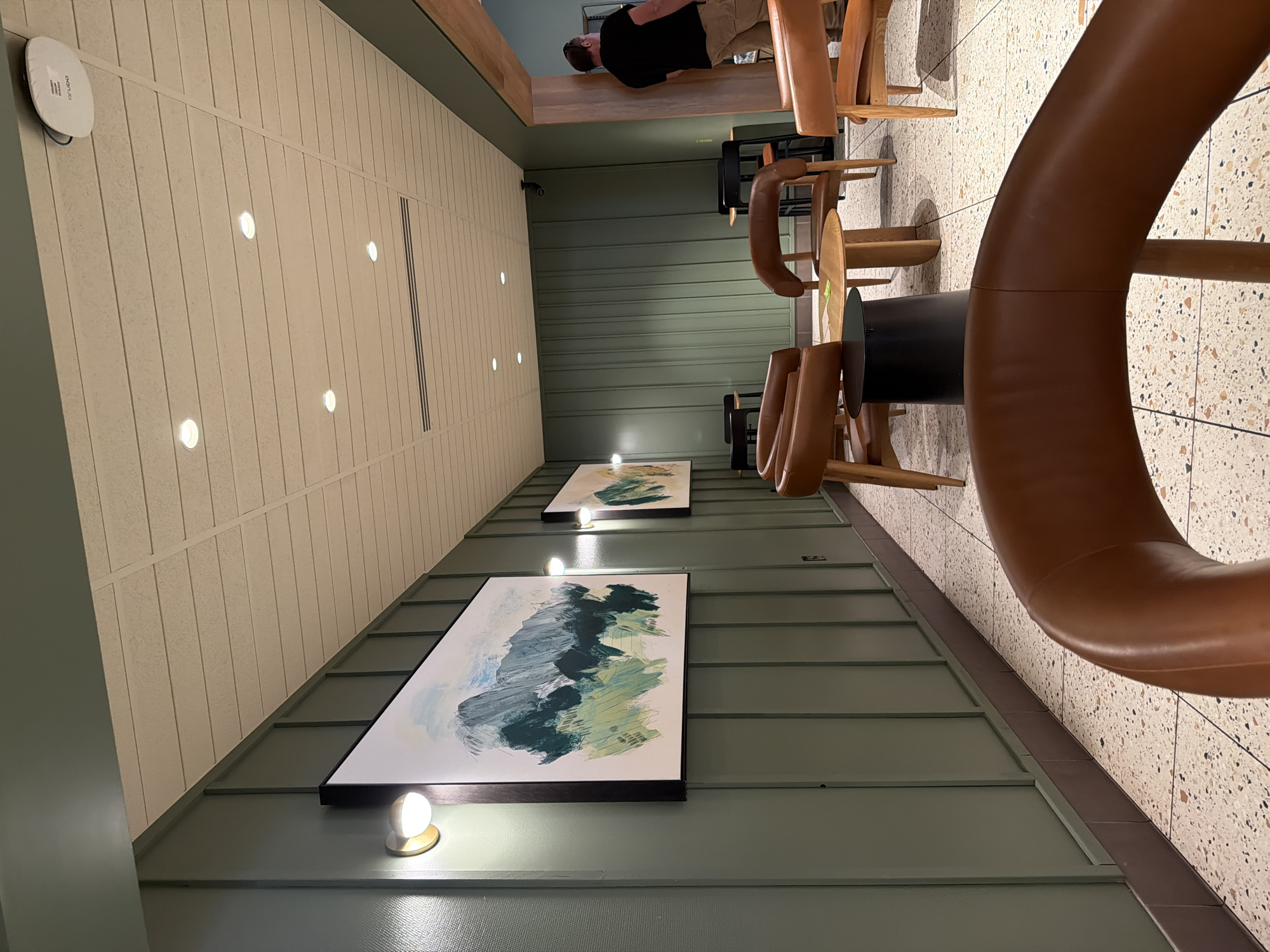 The image depicts the interior of a modern cafe or waiting area in North Bend, United States of America. The space features a distinct design with walls paneled in a muted green color. Several pieces of abstract art, with a focus on blues and greens, are hung on the walls. The seating area is furnished with sleek, modern chairs upholstered in warm brown leather, arranged around small wooden tables. A dark cylindrical side table is also present. The floor is a speckled terrazzo pattern. The ceiling is a neutral tone with inset lighting and what appears to be a Wi-Fi access point mounted on it. Natural light seems to be complemented by artificial lighting from wall-mounted fixtures and overhead spotlights. In the background, a man wearing a dark t-shirt and light-colored pants is seen walking through a doorway into an adjacent area.