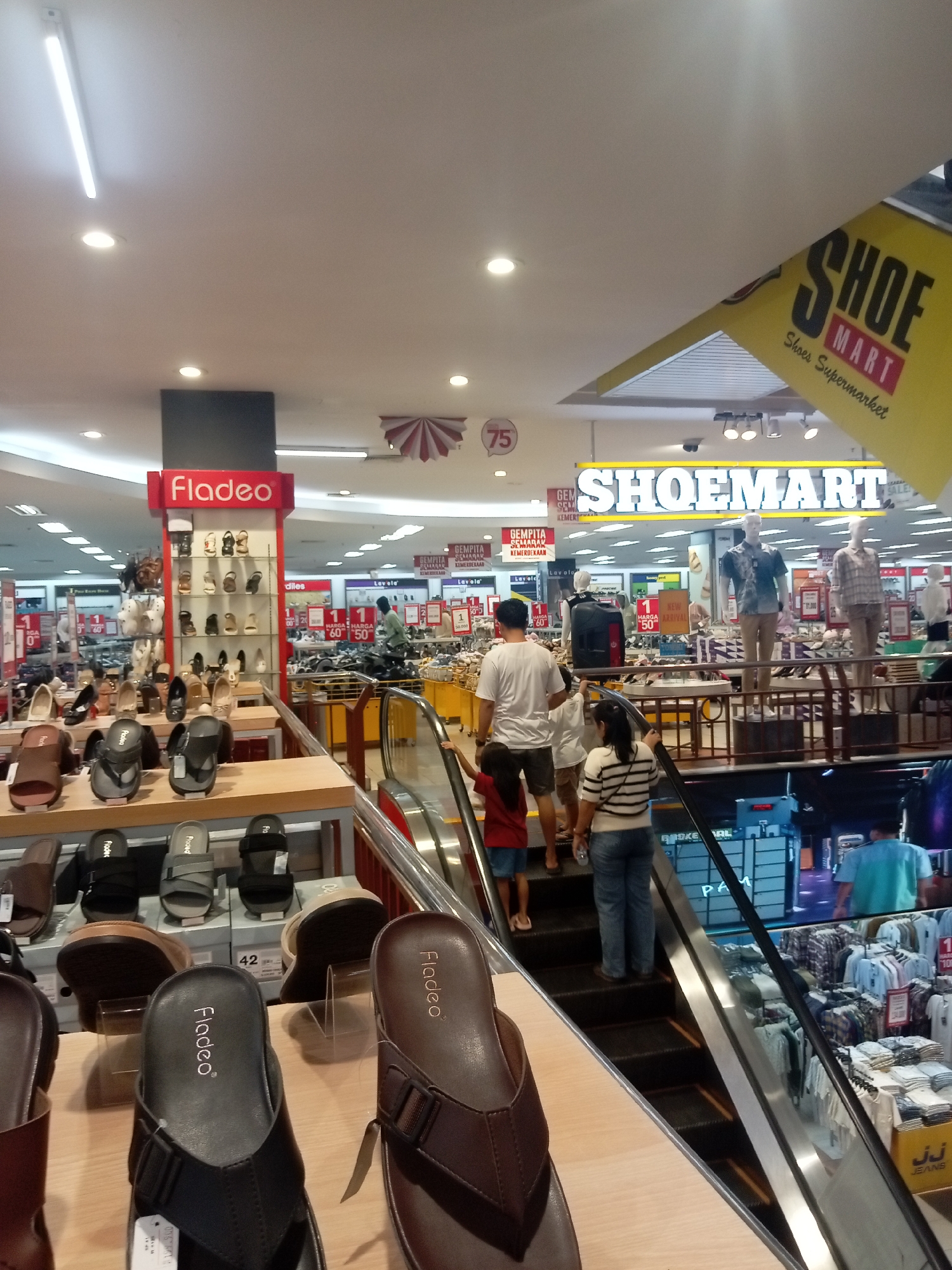 The image shows the interior of a shoe store, likely in Batam, Indonesia, given the signage "Shoe Mart" and "Fladeo" which are common brands in Southeast Asia. The store appears to be a large retail space with multiple displays of footwear and clothing. In the foreground, several pairs of sandals are prominently displayed on wooden shelves. An escalator is visible in the midground, with a family ascending it. The father, wearing a white t-shirt and shorts, is leading the way, followed by his son, daughter, and wife. The wife is wearing a striped t-shirt and jeans. The background reveals more rows of shoes and clothing racks, as well as mannequins dressed in shirts and pants. A large sign with the words "SHOE MART" is prominently displayed overhead. The store is well-lit with fluorescent lighting.