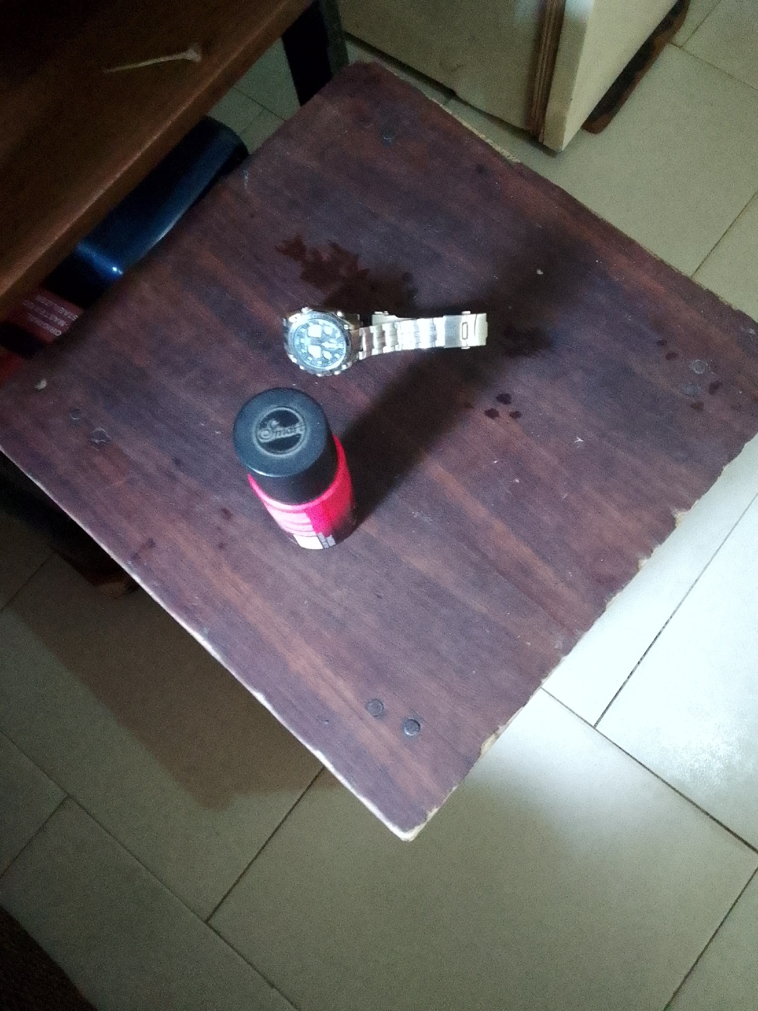An overhead shot shows a dark, wooden table with a watch and a small red bottle on it. The watch, made of silver-colored metal, is positioned to the right of the bottle and displays a circular face. The bottle, with a black cap, is bright red and appears to be a cosmetic product. The table's surface is textured and shows some wear, with a few dark spots that might be stains or watermarks. The background is blurred, but hints at a tiled floor and part of a wooden cabinet. This image appears to be taken indoors in an urban setting, possibly a home or a shop. The overall lighting is dim, casting subtle shadows on the table.