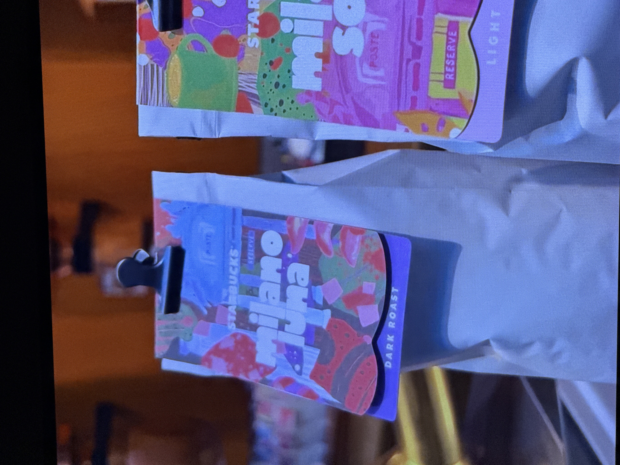 The image shows two coffee bags, one above the other, against a white background. Both bags are decorated with colorful, abstract designs. The top bag has a predominantly purple and pink color scheme with green and yellow accents. Visible text on this bag includes "STAR", "mil", and "RESERVE LIGHT". The bottom bag is also vibrantly decorated with pink and red shapes on a purple background. Text on this bag includes "STARBUCKS", "RESERVE", "JILARA", and "DARK ROAST". A black clip is attached to the left side of the bottom bag. The overall presentation suggests these are retail products, likely coffee beans, displayed for sale or review. The setting is ambiguous, but the clean white background and the focus on the products imply a commercial or display environment. There are no people visible, and the scene is static. The lighting appears to be artificial and even.