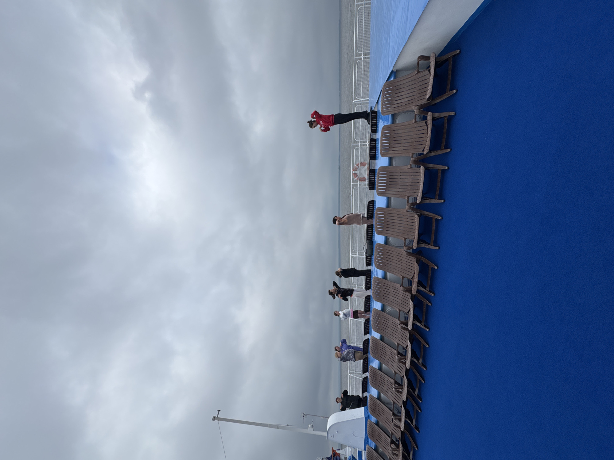 On the deck of a vessel under a cloudy sky, a group of individuals are observed. In the foreground, a row of empty, brown wooden lounge chairs with metal frames are arranged. The deck itself is covered in a vibrant blue material. In the background, several people are standing, some of whom appear to be engaged in stretching or light exercise, with their arms raised or hands placed on their heads. One person in a red hoodie is prominently positioned on a raised platform, also with hands raised. The vessel is at sea, with a calm body of water and a distant shoreline visible under the overcast conditions. The location is identified as Laishevo, Russia.