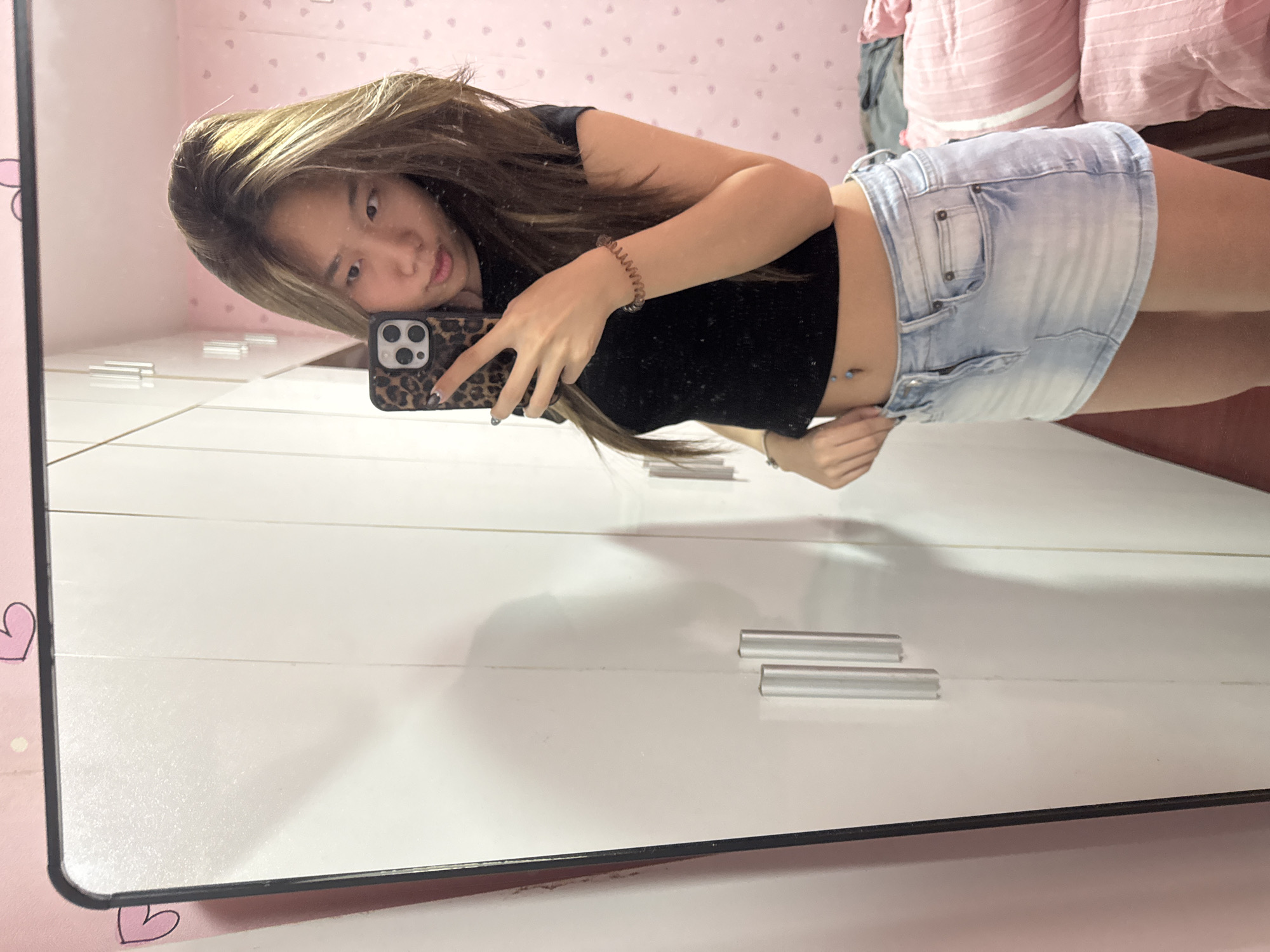 A young woman is taking a selfie in a mirror. She is wearing a black crop top and a short denim skirt. She has long brown hair with blonde highlights. Her phone has a leopard print case. The background includes a white wardrobe with silver handles and a pink wall with heart decorations. The location appears to be an indoor setting, potentially a bedroom. The city of Shapingba District, China, cannot be confirmed from the image alone.