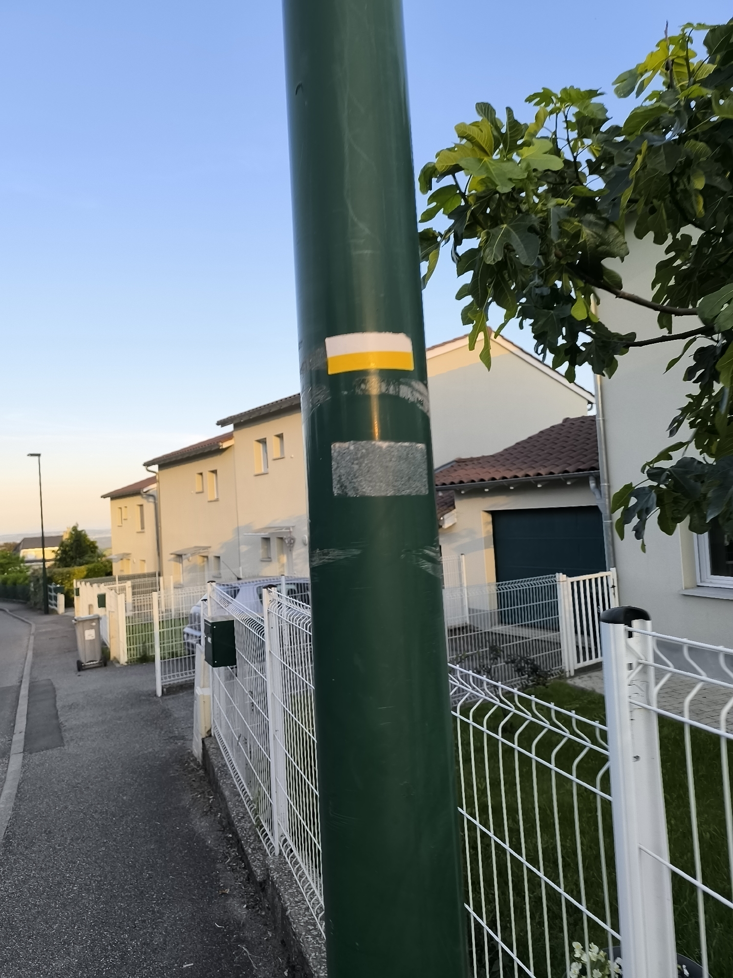 The image shows a residential street in Pélussin, France. A green pole stands in the foreground, with a yellow and white hiking marker affixed to it. Behind the pole, a white metal fence runs along the sidewalk, separating the street from the residential properties. Several houses are visible in the background, painted in light colors with red-tiled roofs. A tree with green leaves is on the right side of the frame. A sidewalk and street run along the left. A trash bin and a lamp post are further in the distance. The sky is light blue.