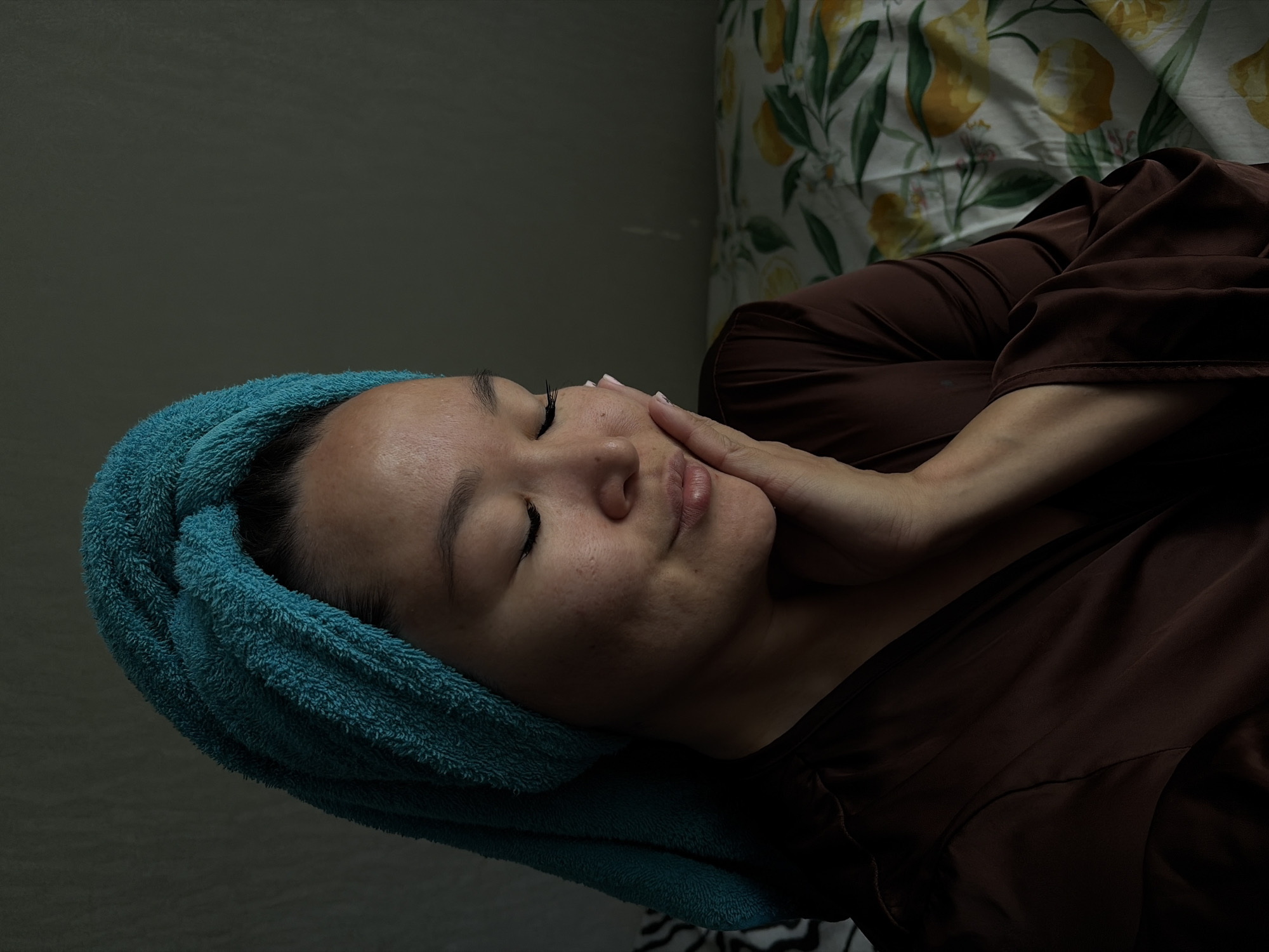 A woman with her eyes closed and hand on her cheek is positioned in the center of the frame, wearing a brown robe and a teal towel turban. She is looking down with a gentle smile. The background includes a wall with a subtle textured pattern and a bed with a yellow and green floral print. The lighting is dim, with a strong highlight on the woman's face and neck. The scene appears to be indoors, possibly a bedroom. The location in Chaikovsky, Russia, is not discernible from the image.