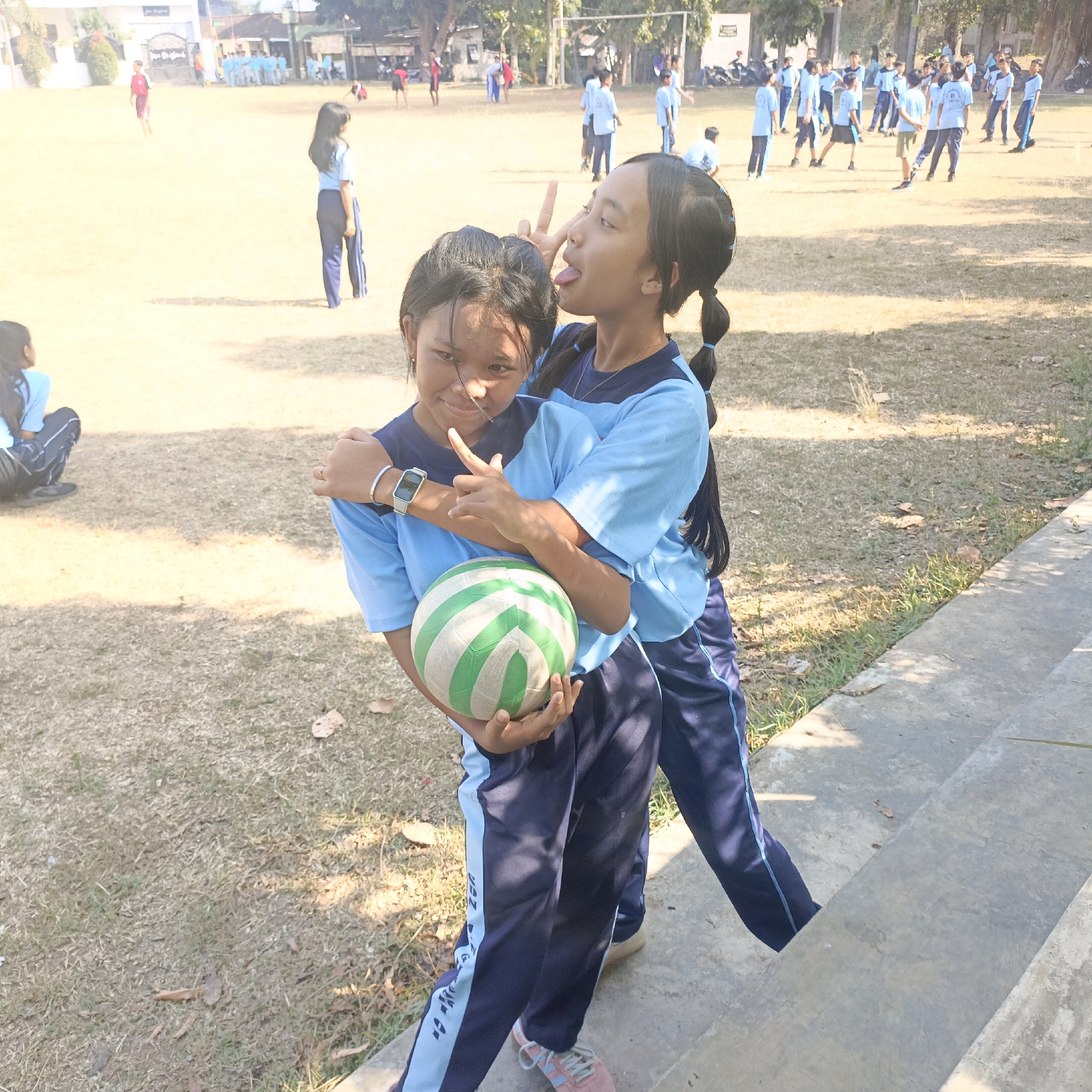 The image depicts two young girls on a sports field, likely during a school event or practice in Salatiga, Indonesia. The girl in the foreground, on the left, is wearing a dark blue and light blue uniform, holding a volleyball. She is looking towards the camera with a slight smile. Behind her, the second girl, also in a similar uniform, is playfully making a peace sign with her fingers and sticking out her tongue towards the camera. She has her arm around the first girl. Both girls have dark hair, one with her hair in a ponytail. The background shows a dry, grassy field with scattered trees and other students in the distance, also in athletic uniforms, suggesting a sports activity. The lighting indicates it is daytime, likely under bright sunlight, creating shadows on the ground. A concrete structure, possibly bleachers or steps, is visible on the right side of the image.