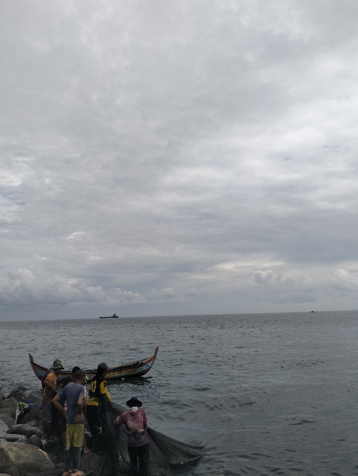 A group of people stands by a fishing boat along a rocky shoreline under a cloudy sky. They appear to be pulling a fishing net from the water. A ship is visible on the horizon. The scene is overcast, with a gray ocean and sky. The group is clustered near the boat, engaged in their fishing activity.