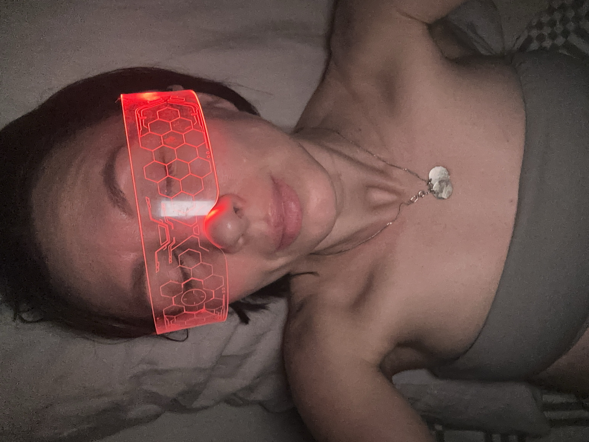 A woman lies on a bed, wearing futuristic red glasses that illuminate with a geometric pattern. She also wears a simple necklace. The background includes bedding and a glimpse of a patterned surface. There is no indication of Votkinsk, Russia.
