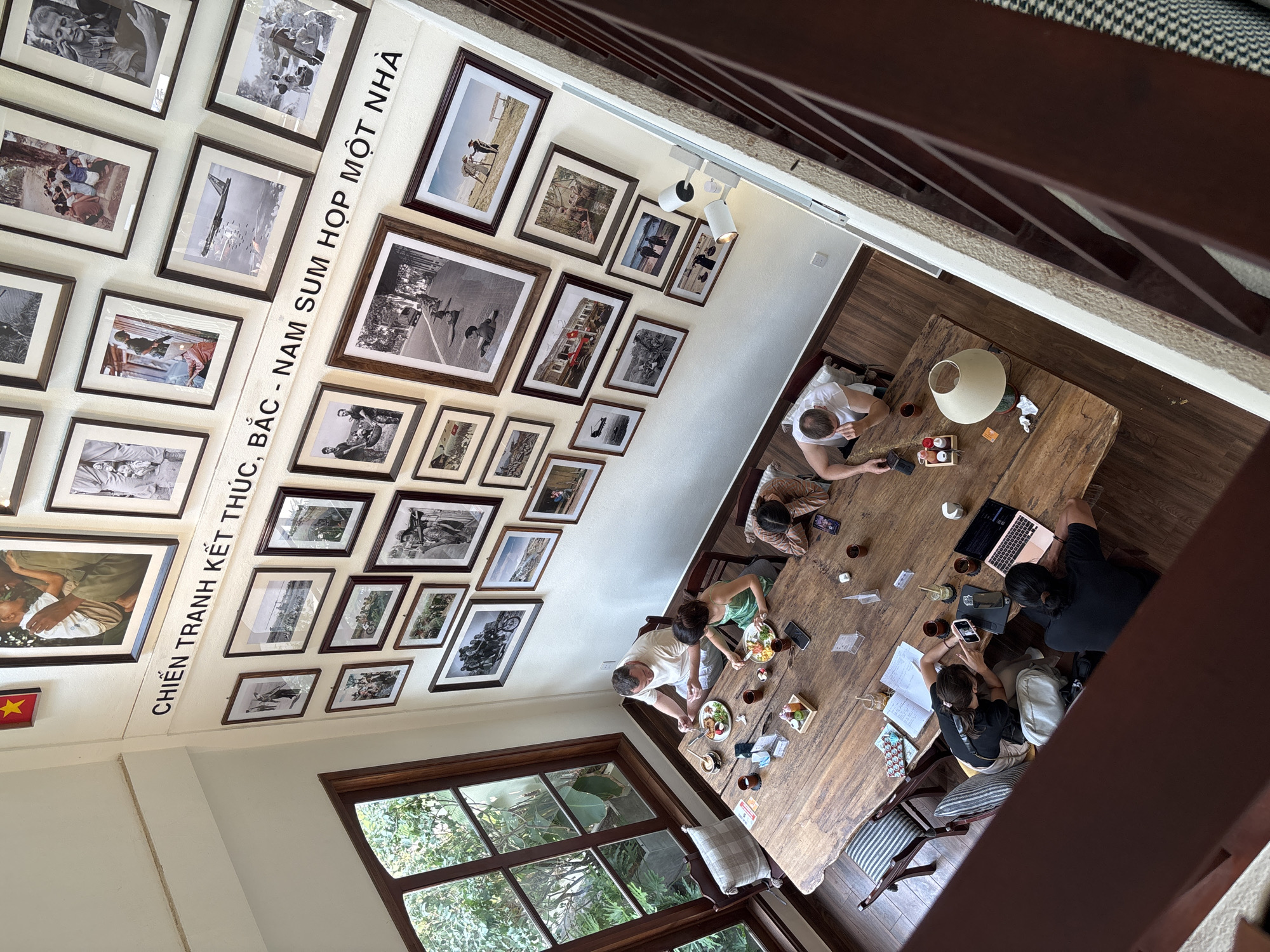 This is an indoor shot taken from above, likely a balcony, showing a large wooden table with a group of people. There are seven people seated around the table, engaged in various activities, including eating, reading, and using electronic devices like phones and laptops. The table is cluttered with dishes, cups, notebooks, and other personal items.
The room has a high ceiling, and one wall is adorned with a series of framed black-and-white photographs. A white banner with black text runs horizontally across the wall amidst the frames. A large window with a wooden frame allows natural light to enter the room, revealing a view of lush greenery outside. The room has a casual and relaxed atmosphere. Based on the text on the wall "CHIẾN TRANH KẾT THÚC - BẮC - NAM SUM HỌP MỘT NHÀ" and the architectural style, this photograph was likely taken at a cafe or restaurant in Đà Nẵng, Vietnam.