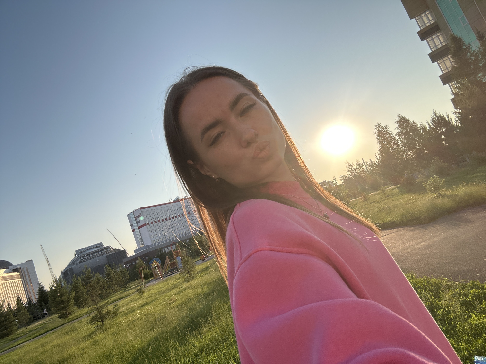 A woman is taking a selfie in a park with buildings and trees in the background. The woman is wearing a pink sweatshirt and has her lips puckered. The sun is setting, casting a warm glow on the scene. The park appears to be well-maintained with green grass and trees. In the background, there are several modern buildings, some of which appear to be under construction, along with a playground. The overall atmosphere is bright and cheerful.