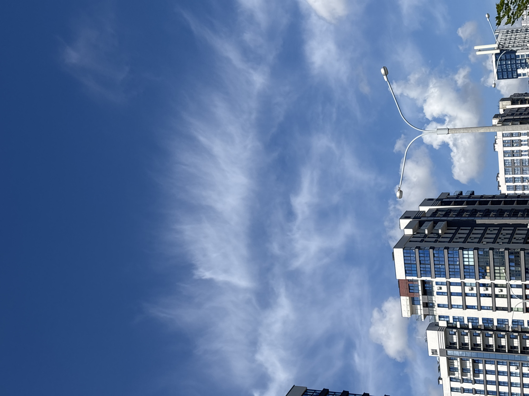 The image captures a clear blue sky filled with wispy cirrus clouds over a modern urban landscape in Minsk, Belarus. In the foreground, several multi-story apartment buildings with contemporary architecture are visible. These buildings feature a mix of white and dark grey facades, with numerous balconies and large, reflective windows. The buildings are densely packed, suggesting a residential district. A prominent street light with two lamps stands centrally in the mid-ground, its white poles curving upwards against the sky. Other street lights and elements of urban infrastructure, such as antennas on a building, are also present. The scene appears to be devoid of people. The overall impression is that of a bright, sunny day in a developed area of Minsk.