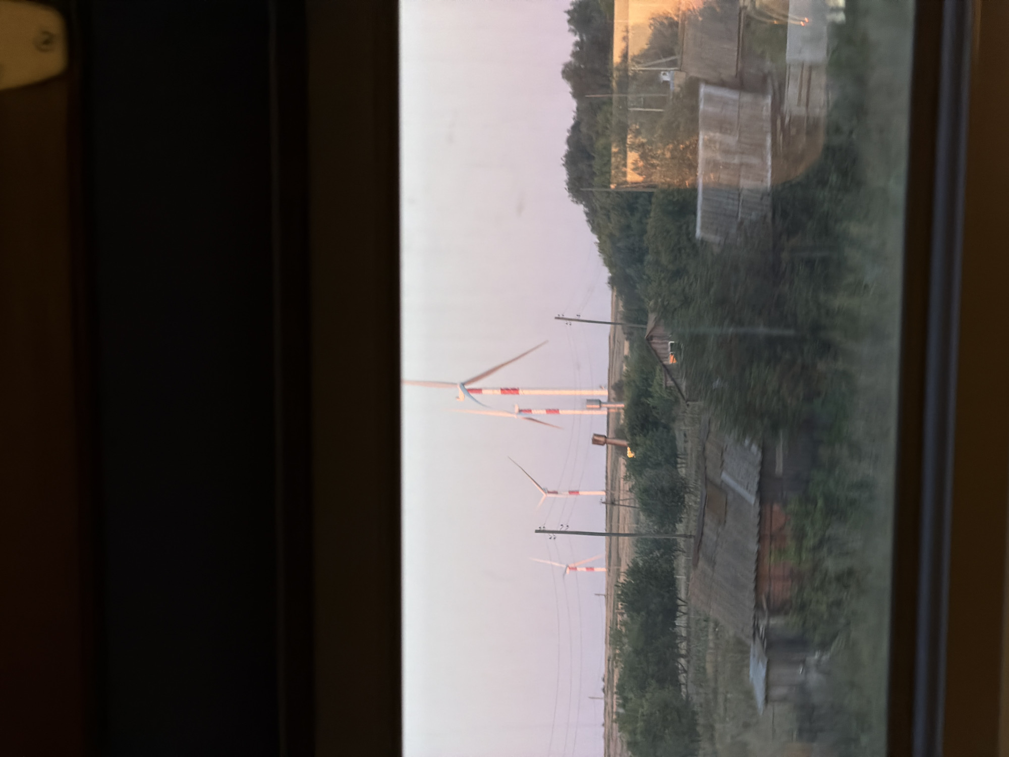 The image depicts a rural landscape in the vicinity of Karamysh, Russia, viewed through a window. In the background, several modern wind turbines with white and red striped towers are visible against a pale pink sky, suggesting either dawn or dusk. The midground is characterized by a flat, grassy field dotted with trees and power lines. Several rural structures, appearing to be houses or sheds with corrugated roofs, are scattered across the landscape. One prominent structure in the foreground on the left is a wooden house with a weathered roof. The overall impression is one of a sparsely populated, agricultural area with renewable energy infrastructure. The photograph is taken from inside a moving vehicle, indicated by the slight blur and reflections on the window.