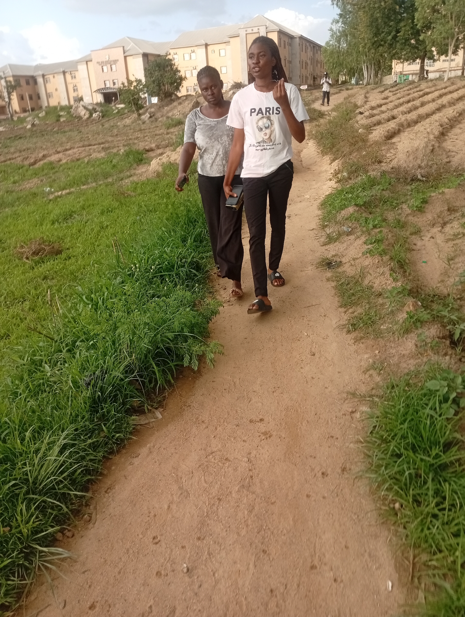 Two young women are walking on a dirt path in Jos, Nigeria. The woman in the foreground is wearing a white t-shirt with the word "PARIS" printed on it in black letters, along with a graphic of sunglasses and a face. She is also wearing black pants and black slides. The woman behind her is wearing a grey t-shirt and black pants. Both women appear to be in their late teens or early twenties.
The path is surrounded by green grass and dry, tilled earth, suggesting an agricultural area or a developing landscape. In the background, there are large, multi-story buildings, possibly dormitories or administrative blocks, set on a hill. Trees are also visible in the background. The sky is overcast, indicating a cloudy day. The overall impression is of a casual walk on a campus or in a semi-urban environment. The subjects do not display strong emotions, appearing to be engaged in conversation or simply enjoying a walk. No other specific activities are evident. The time of day appears to be late afternoon or early evening, given the diffused light.