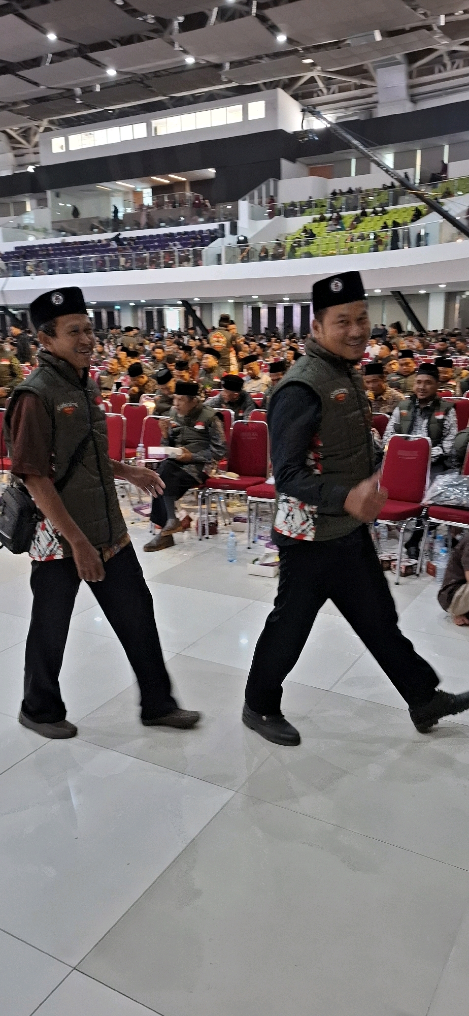 The image depicts an indoor event space with tiered seating and a large crowd of people. Two men in the foreground, wearing black caps and olive-green vests over patterned shirts and black pants, are walking. The man on the left, with a darker complexion, smiles and gestures with his left hand. The man on the right, with a lighter complexion, also smiles and gives a thumbs-up with his right hand. The background reveals a large audience seated in red chairs, many wearing similar attire to the men in the foreground. The environment appears to be a modern, spacious hall with a high ceiling and lighting fixtures. The location is identified as Blulukan, Indonesia, suggesting this event is taking place there.
