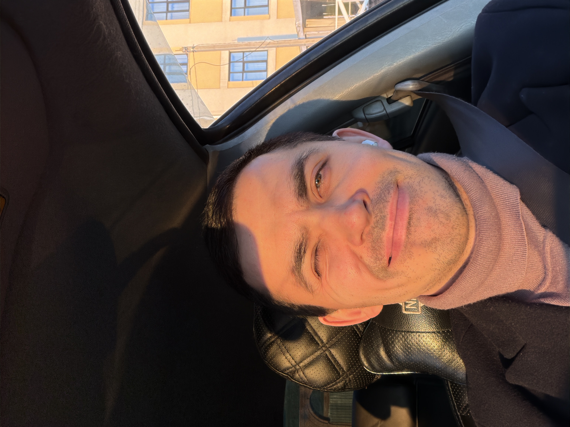 A man is captured in a selfie or portrait, seated in the passenger or driver's seat of a car. The image is oriented with the man's head facing the right side of the frame, giving a rotated view. He is smiling and winking with his left eye, conveying a playful and relaxed demeanor. Strong, warm sunlight illuminates the right side of his face and parts of the car's interior, indicating either early morning or late afternoon light (golden hour). He has short dark hair, light stubble on his chin and cheeks, and a light-colored earbud in his right ear.
He is wearing a light pink turtleneck sweater under a dark-colored jacket or coat. A dark seatbelt is visible across his chest and shoulder. The car's interior features a black leather seat with a distinctive quilted pattern on the headrest. Partially visible on the headrest is a white logo or text, possibly starting with "N".
Through the car window, a portion of a building is visible outside. The building is a light yellow or ochre color with multiple rectangular windows, some appearing blue-tinted, suggesting a sunny day. The background elements imply an urban or suburban setting, consistent with Izhevsk, Russia, given the provided context. The overall scene suggests a moment of leisure or travel, captured with good natural lighting.