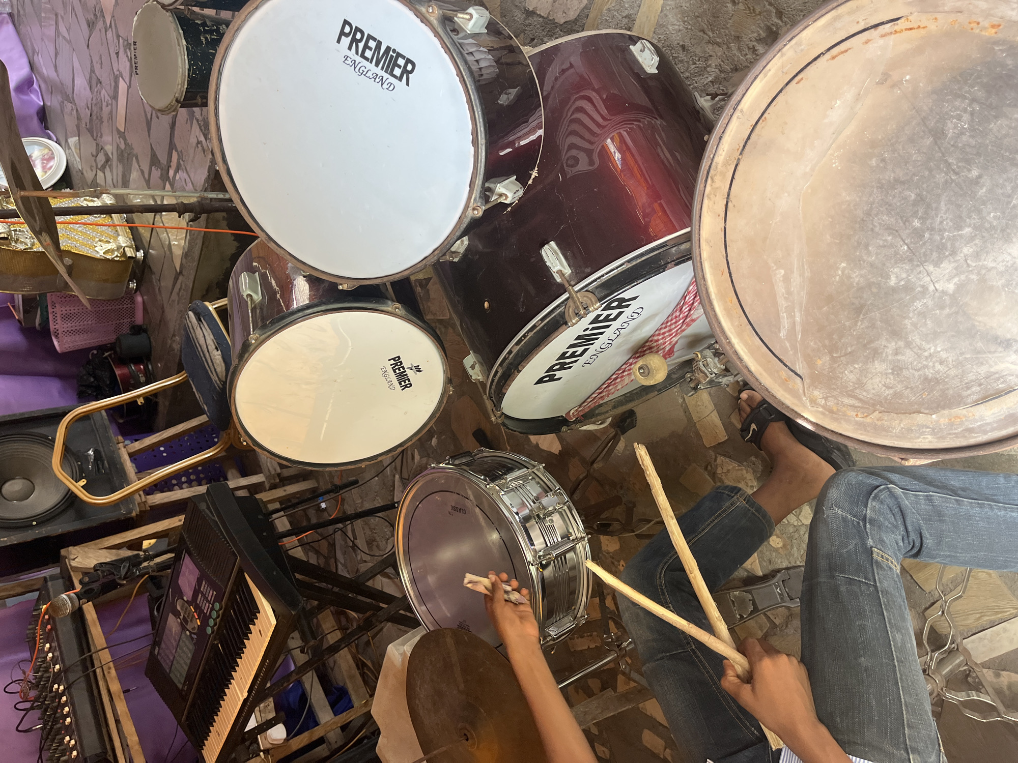 The image depicts a set of Premier brand drums located in Iranla, Nigeria. The drum set includes a bass drum, snare drum, and several tom drums. A man wearing jeans is seated behind the drums and holding drumsticks. A keyboard, speaker, and a microphone with a mixer are visible to the left. The location appears to be an indoor space with a purple cloth partially visible in the background.