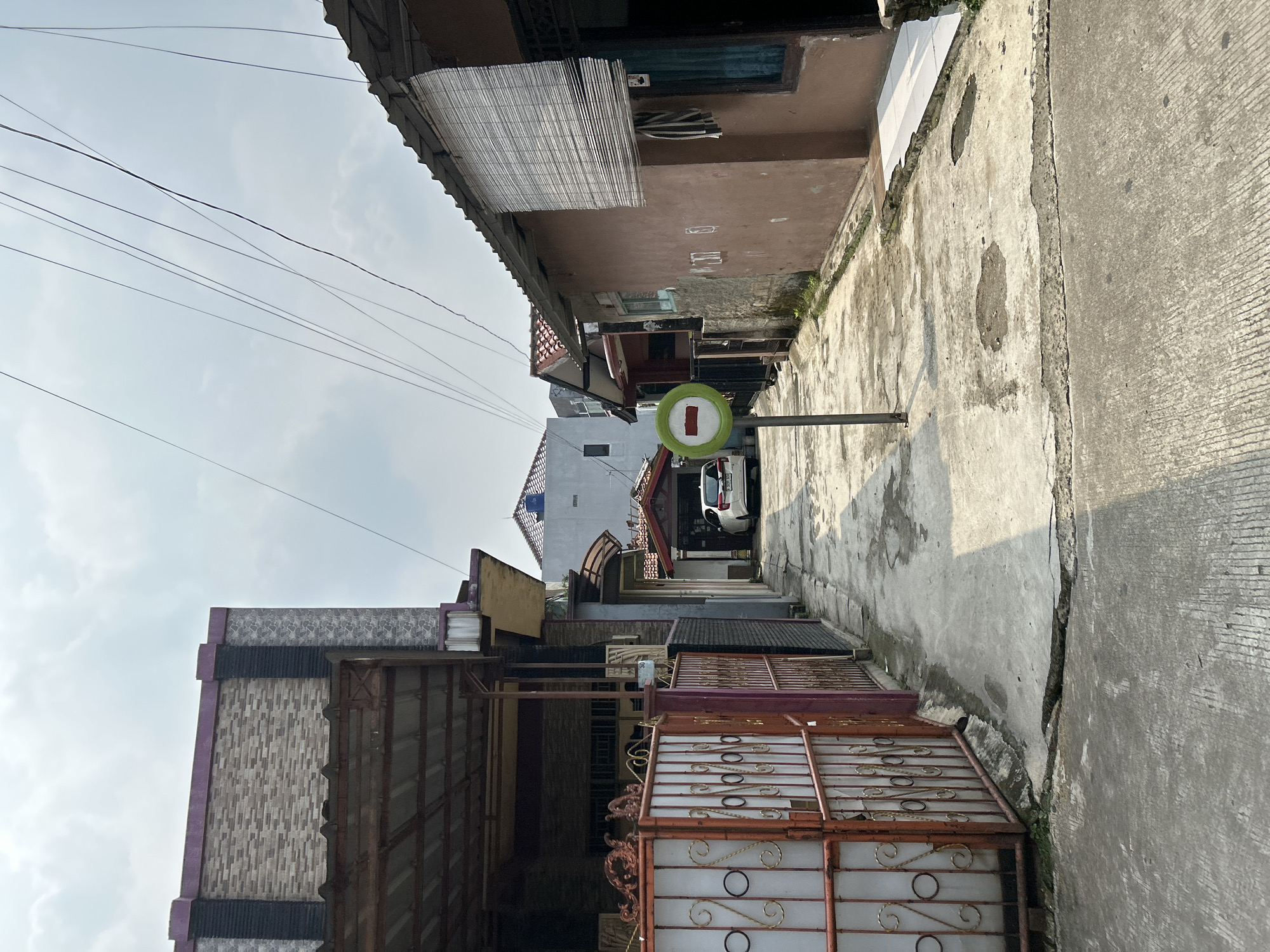 The image depicts a narrow, paved alleyway in a residential area, likely in Depok, Indonesia. The alley is flanked by houses with a mixture of architectural styles and building materials. On the left, a property features a tall, ornate red metal gate with decorative swirls and circular motifs, leading to a covered carport. The building behind the gate has a textured brick facade and a dark-colored roof. The alleyway itself is made of concrete, showing signs of wear and tear with cracks and patches.
In the center of the alley, a circular traffic sign with a red border and a white horizontal bar signifies a "No Entry" or "Do Not Enter" restriction. A white car is parked further down the alley, partially obscured by the sign. The houses on the right side of the alley have various roofing materials, including corrugated metal and red tiles. One house has a brown-colored facade with a woven bamboo blind covering a window. Overhead, power lines are strung across the sky, contributing to the urban feel of the scene. The sky is partly cloudy.