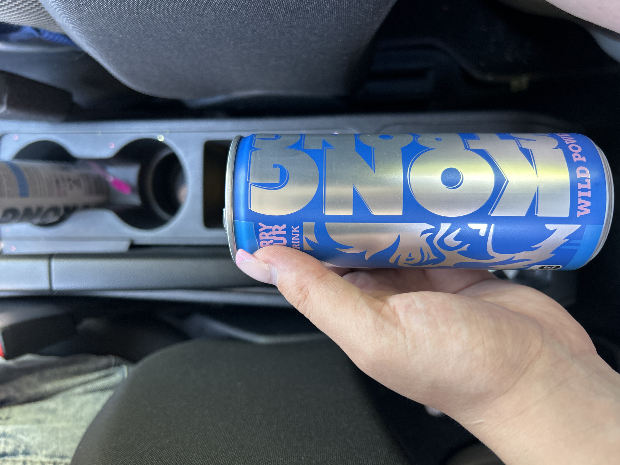 A person's hand is holding a blue and silver can of "KOONG WILD POWER" energy drink. The can has a graphic of a roaring lion or tiger on it and text that reads "BERRY SOUR DRINK". The can is positioned in the foreground, with a car's center console and seats visible in the background. The center console has two cup holders, one of which contains another can. The scene appears to be inside a vehicle, possibly in the city of Reichshof, Germany, given the provided context. The lighting suggests it is daytime.