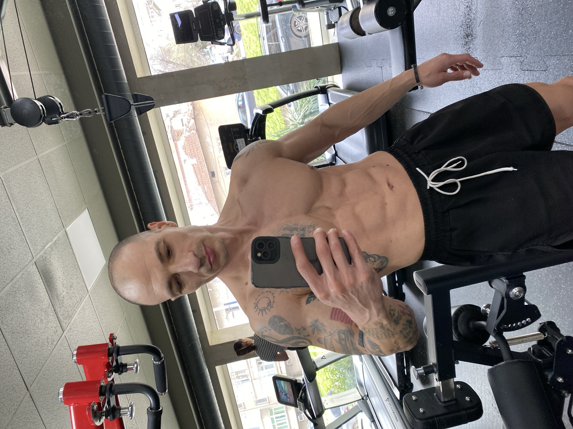 The image depicts the interior of a gym in Pleven, Bulgaria. A light-skinned adult male, shirtless and with a shaved head, stands in the foreground, holding a dark-colored smartphone in his right hand, pointed towards himself, indicating he is taking a selfie. He wears black shorts with a white drawstring and has extensive tattoos on both arms, including a sun and crescent moon motif and a microphone on his right bicep, and various designs on his left arm. A black bracelet is visible on his left wrist. His physique shows visible musculature, and a surgical scar is present near his navel.
In the mid-ground, to the left, a light-skinned adult female with brown hair tied back is seen from behind, wearing a striped shirt, walking on a treadmill. The gym features various equipment, including red and black weight machines, multiple treadmills with digital displays, and dumbbells on a rack. The floor is covered with grey interlocking rubber mats. Overhead, a textured acoustic tile ceiling is visible, along with exposed black ventilation ducts and a black cable pulley system. Large windows line the back wall, revealing an exterior urban environment with residential buildings, some featuring reddish-brown tiled roofs, parked vehicles, and sparse greenery under natural lighting conditions.