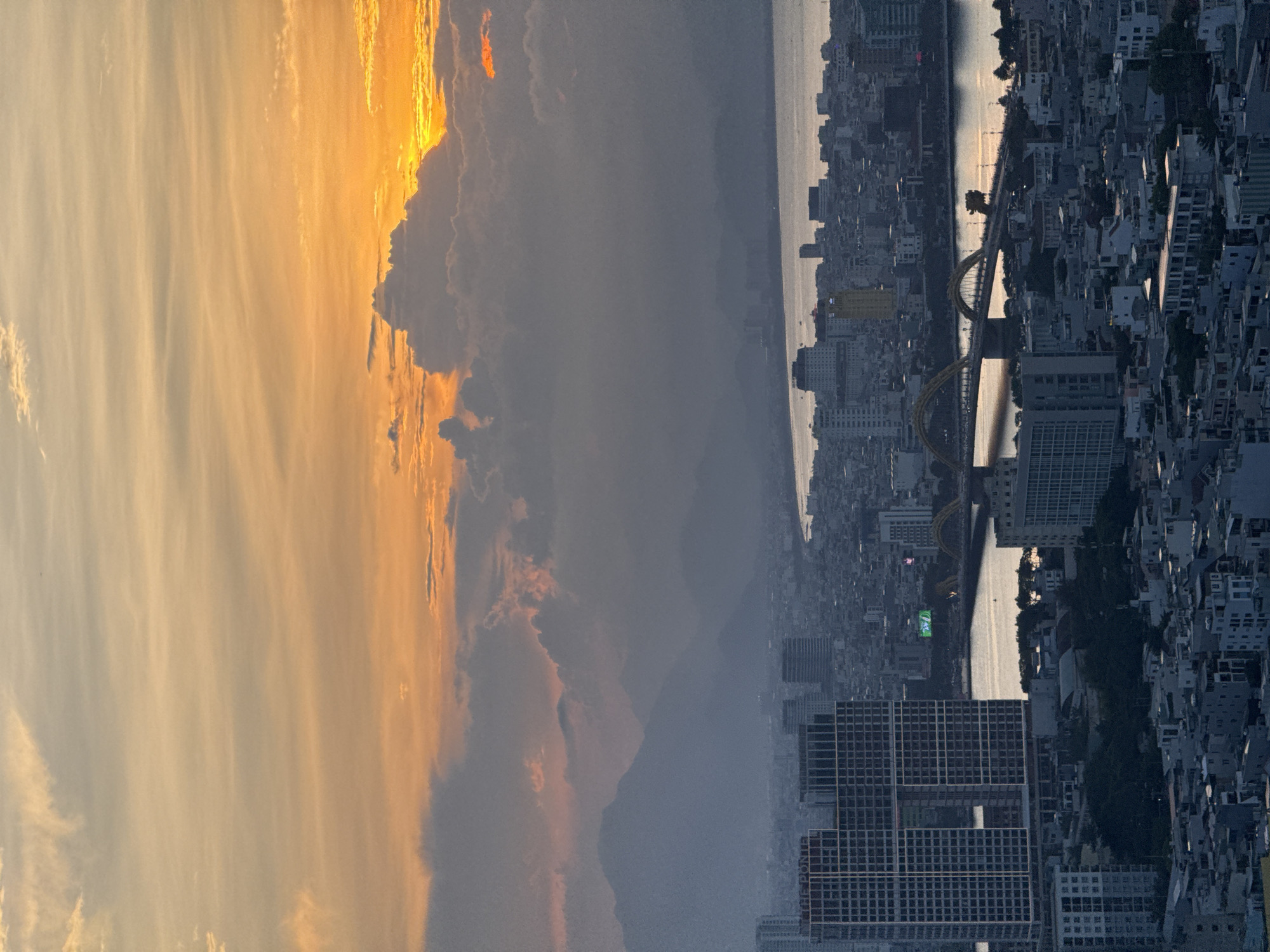 This is an aerial shot of a city during sunset. The sky is filled with soft orange and yellow hues from the setting sun, peeking through scattered clouds. In the foreground, there are numerous buildings, some tall and modern, others smaller and densely packed. A prominent river cuts through the city, spanned by a distinctive yellow bridge with an arched design. In the background, silhouetted mountains rise against the colorful sky, shrouded in a hazy mist. The city appears to be located in a coastal area, as the river flows towards a body of water on the right side of the image. The lighting suggests it is either early morning or late evening. The specific architectural style of the buildings, particularly the large grid-like facade of the building labeled "THE PONTE" and the presence of the Dragon Bridge, strongly suggest the location is the city of Đà Nẵng, Vietnam. There are no discernible people in the image.