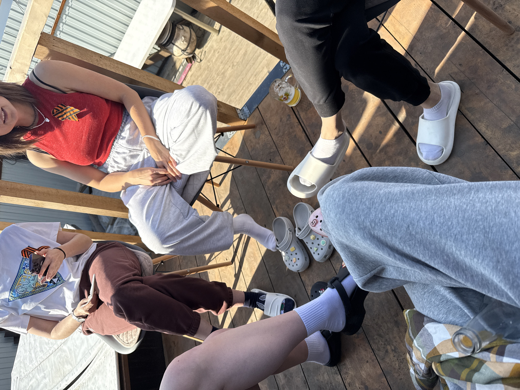 Four people sit on chairs on a wooden platform. The person on the left wears a white t-shirt and brown pants, holding a phone. Next to them is someone wearing a red tank top and grey sweatpants. To the right is a person in black pants. Another person wears gray sweatpants. A glass of beer sits on the platform. Several pairs of Crocs and slides rest on the wooden surface. The location is most likely Krasnoyarsk, Russia, indicated by imagery on the clothing.