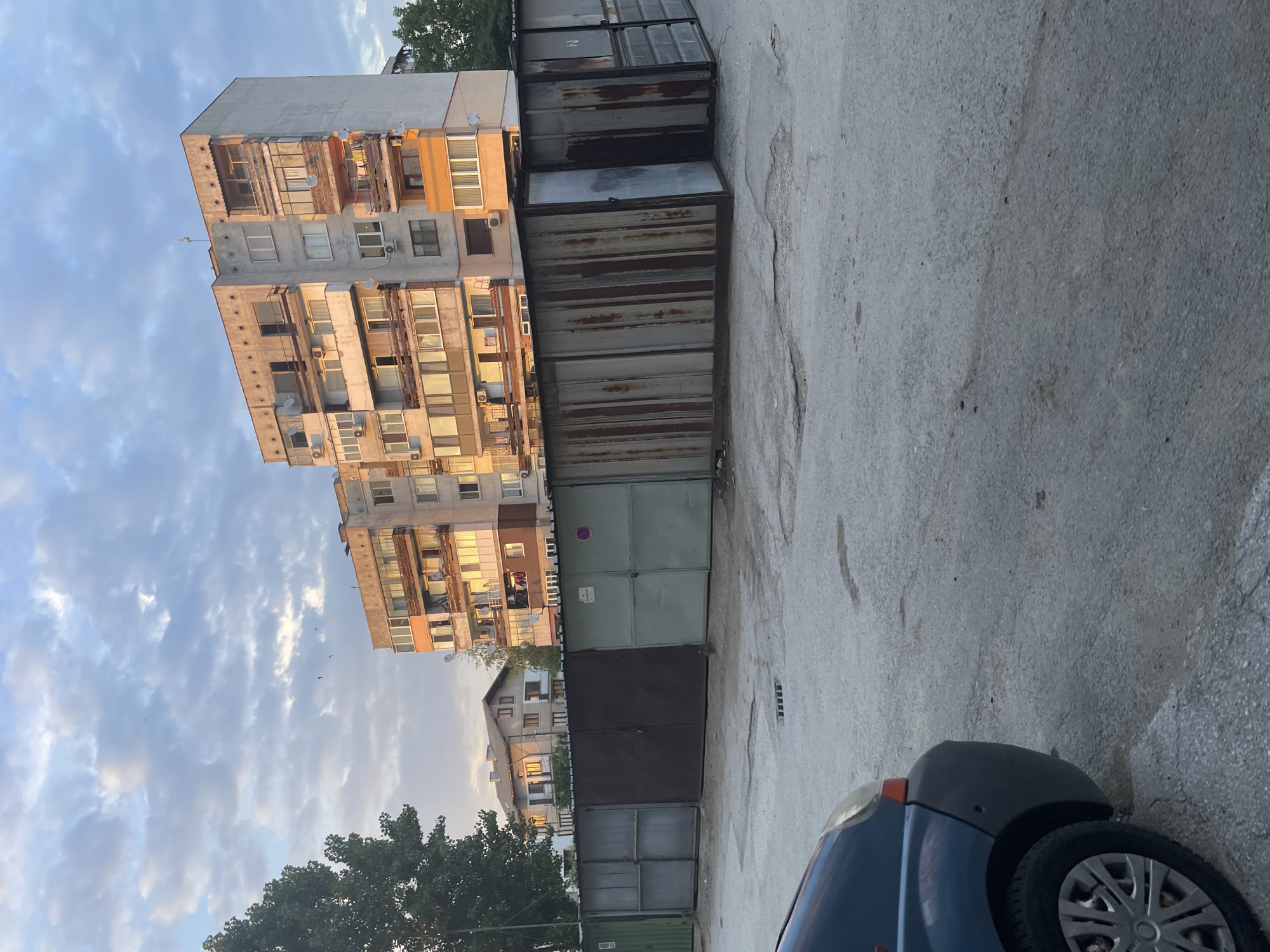 An outdoor scene in Ihtiman, Bulgaria, captured during late afternoon or early morning light, characterized by warm, directional illumination. The foreground features an uneven asphalt or concrete surface with minor cracks and stains, containing a small metal grate. A partial view of a dark-colored vehicle, likely a van or hatchback, with a silver hubcap, is visible on the far left. Stretching across the mid-ground is a linear arrangement of approximately ten single-story garages or storage units, displaying varied states of repair and materials; some are dark grey, dark brown, or light green, while others are corrugated metal, extensively rusted, with vertical seams. One light-colored garage door displays a sticker and a red "no parking" sign.
In the background, two multi-story residential apartment buildings with concrete facades dominate the scene. These structures exhibit multiple balconies, some enclosed, others open with diverse railings. Numerous satellite dishes and external air conditioning units are affixed to their exteriors. Portions of the buildings, particularly balconies and lower sections, are painted in shades of orange and peach, reflecting the warm sunlight. Behind the garages and to the left of the apartment blocks, a two-story house with a brown roof and light-colored walls is partially visible, also illuminated by the low sun. Trees and green foliage are present on the far left and intermittently between the buildings. The sky above is partially overcast with white and grey clouds, displaying patches of blue, and several small birds are discernible in flight. No individuals are visible.