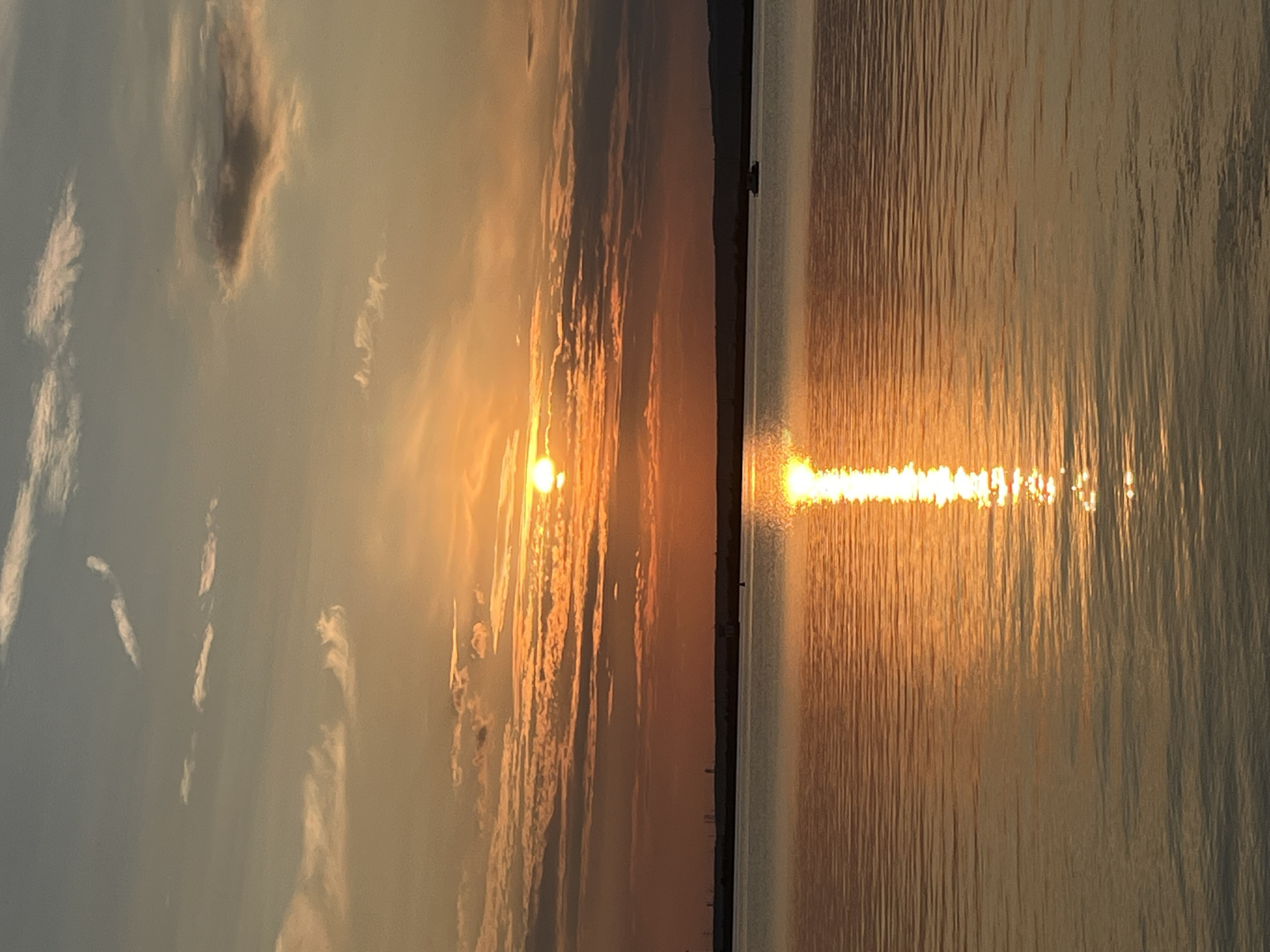 A picturesque sunset over the Kama River in Chaikovsky, Russia, casts a warm orange glow across the sky and reflects vibrantly on the water's surface. The sun, a bright orb, is partially obscured by wispy clouds, creating a dramatic and colorful display. The calm river stretches to the horizon, where a silhouetted landmass is visible. A small boat is seen in the distance on the water. The overall scene evokes a sense of tranquility and natural beauty.