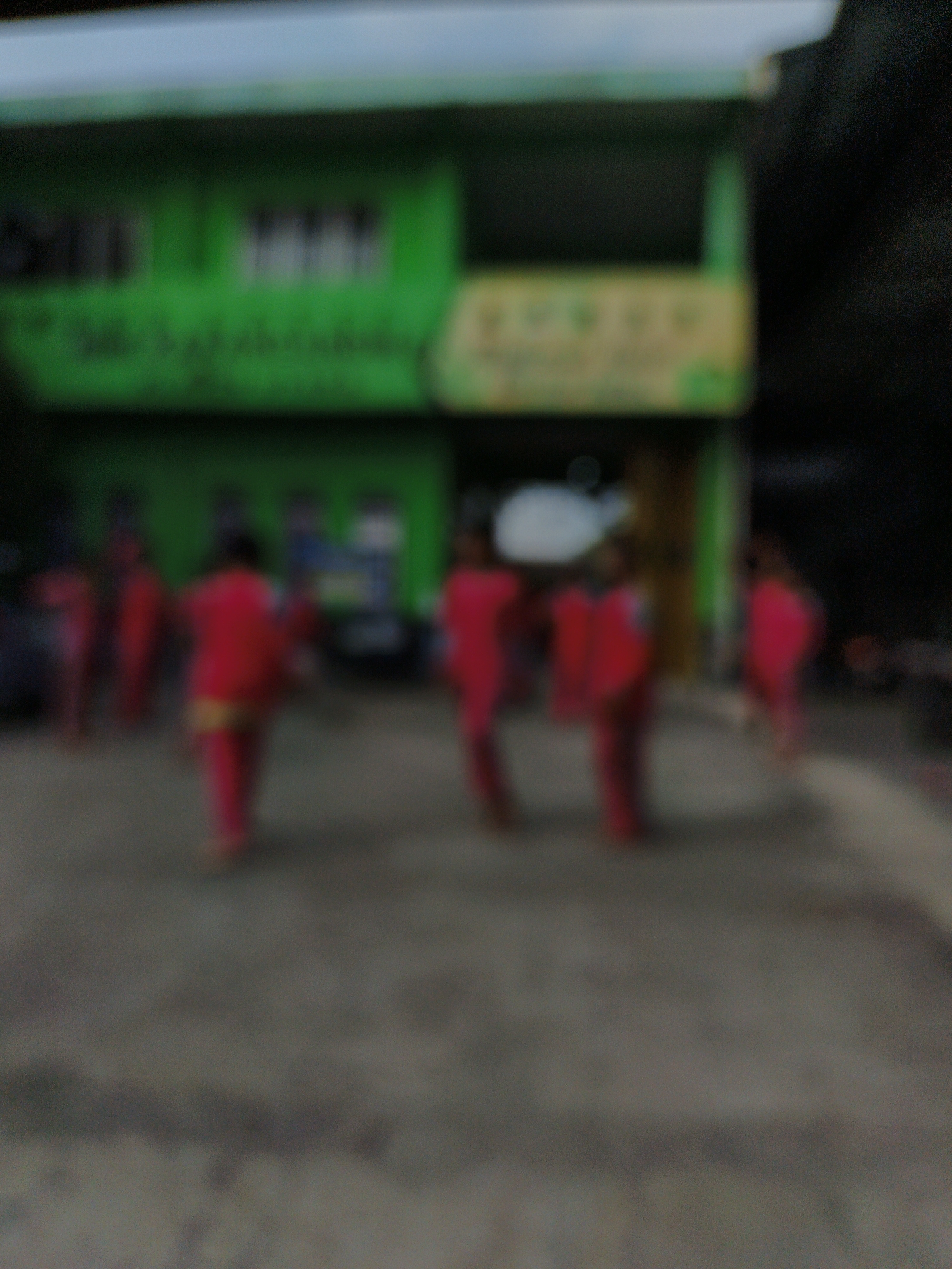 The image, captured in Jonggrangan, Indonesia, is a blurred outdoor scene in front of a two-story green building. Several individuals, appearing to be children, are visible in the foreground, all wearing vibrant pink or red clothing, some with yellow accents. They are positioned on a paved surface, possibly a courtyard or street, in front of the building. The building itself has a light-colored roof and a green facade with some indistinct markings or signage. The overall lighting suggests it might be daytime, though the blurriness of the image makes precise details difficult to ascertain.