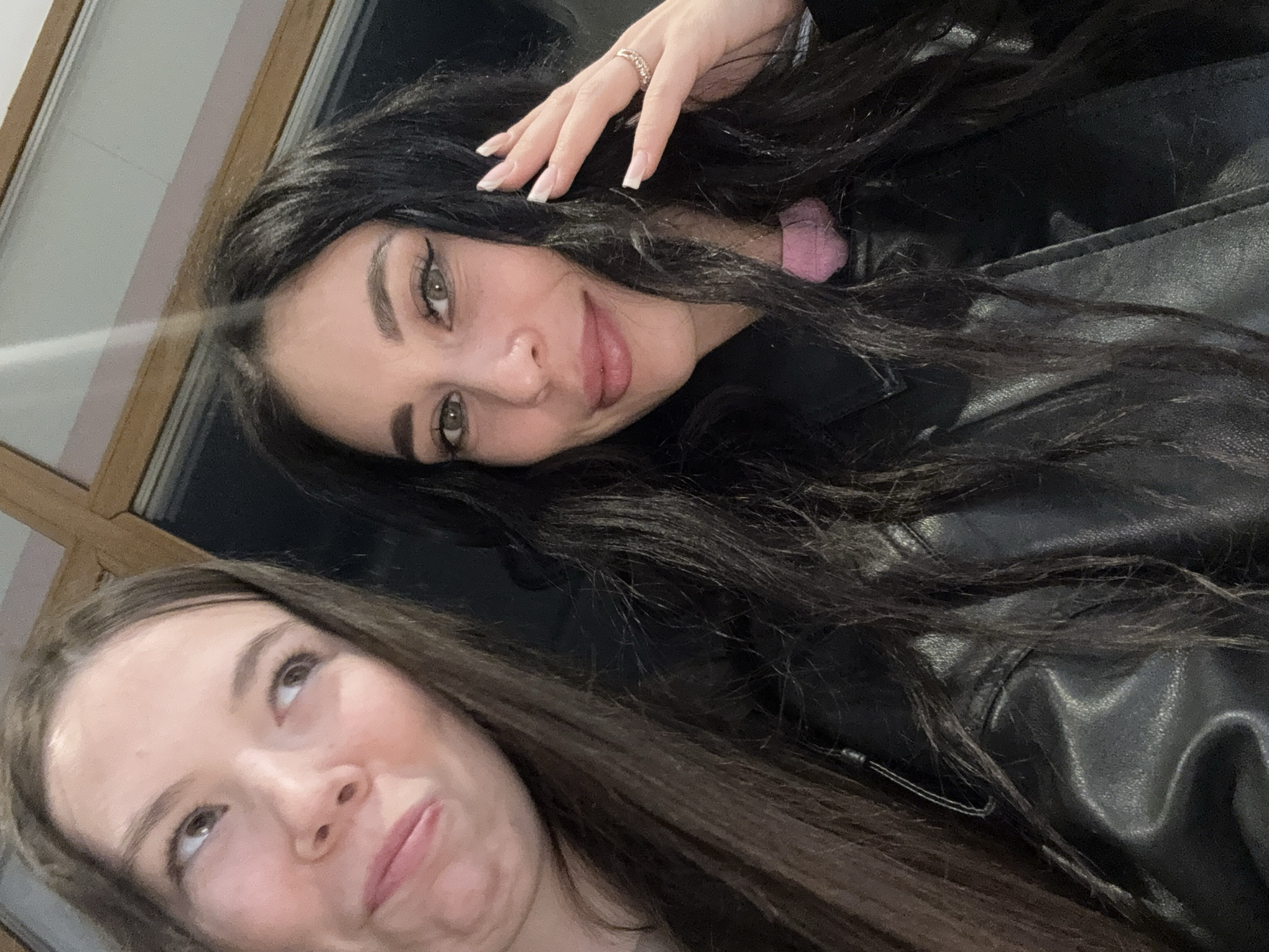 Two women are posing for a photograph. The woman at the top has dark hair, fair skin, and wears a ring on her finger. She is wearing a black leather jacket. The woman at the bottom has light brown hair and fair skin. The photo appears to be taken indoors, possibly near a window or reflective surface. There is no verifiable evidence in the image to suggest the location is in the city of Chaikovsky, Russia.