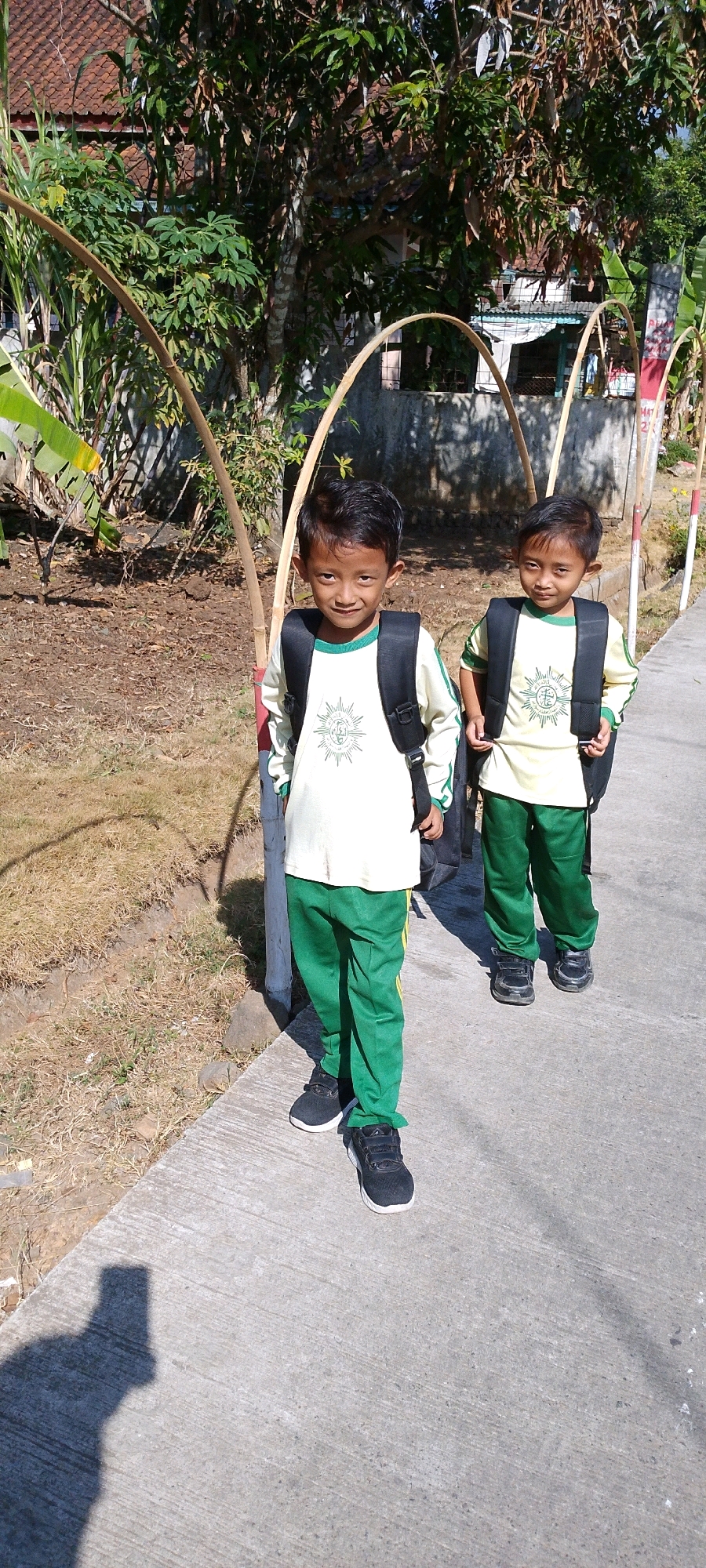 In Sindangrasa, Indonesia, two young boys stand on a concrete path, facing forward. They are dressed in matching uniforms: cream-colored long-sleeved shirts with green trim and green pants with yellow stripes down the sides. Both boys wear black backpacks. The boy on the left is slightly closer to the camera and looking directly at the viewer with a slight smile. The boy on the right is standing a few steps behind and to the right of the first boy, also looking forward.
The background features a dirt and dry grass area to the left, with some scattered vegetation and trees. The path they are standing on is made of concrete and appears to be a sidewalk. Behind them, a concrete wall is visible, topped with greenery and a tiled roof structure. Several arched bamboo structures are positioned along the path, arching over it from the grassy area. The scene is illuminated by bright sunlight, casting shadows. The overall environment suggests an outdoor setting, possibly near a school or residential area.