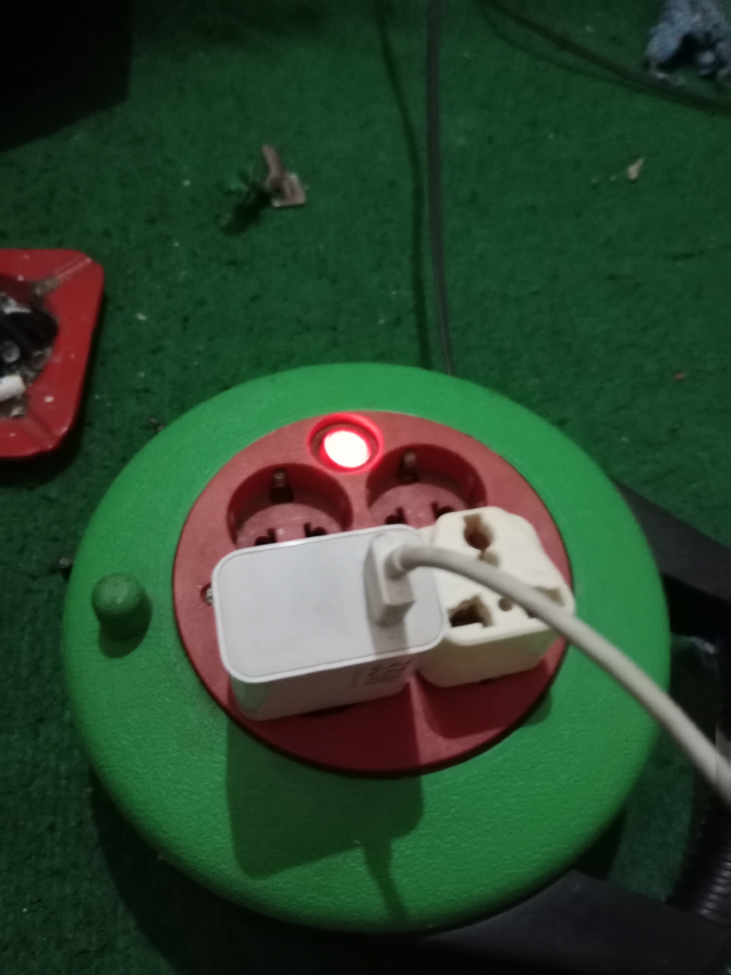 In Ringinrejo, Indonesia, a close-up shot captures a green and red circular power strip resting on a green carpeted surface. The power strip has three visible outlets, two of which are occupied by white electrical adapters. A red indicator light on the power strip is illuminated, suggesting it is active. To the left, a red ashtray containing cigarette butts and a lighter is partially visible. The background is dark and out of focus, with a hint of a black cable extending upwards. The scene appears to be indoors.