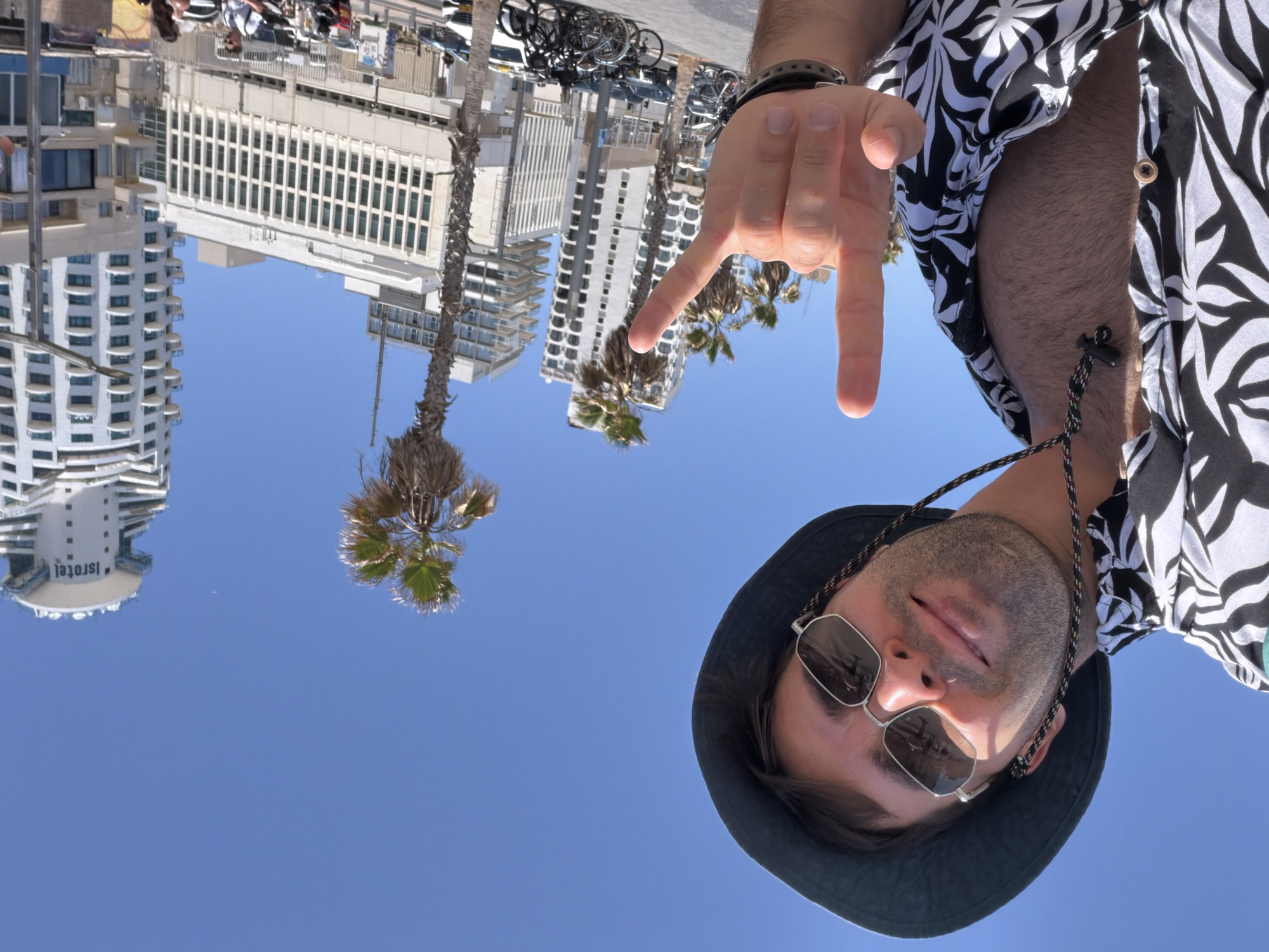 The image is an upside-down selfie taken from a low angle, featuring a person wearing a black bucket hat and sunglasses, with a black-and-white patterned shirt partially visible. The person is making a hand gesture, specifically "rock on." The background reveals a cityscape with modern, white buildings and palm trees, indicating an urban beach environment. The buildings are typical of the beachfront hotels and apartments in Tel Aviv, Israel. The clear blue sky suggests a sunny day. Bicycles and people can be seen in the distant background. The overall composition suggests a casual, tourist-oriented snapshot in a popular location of Tel Aviv.