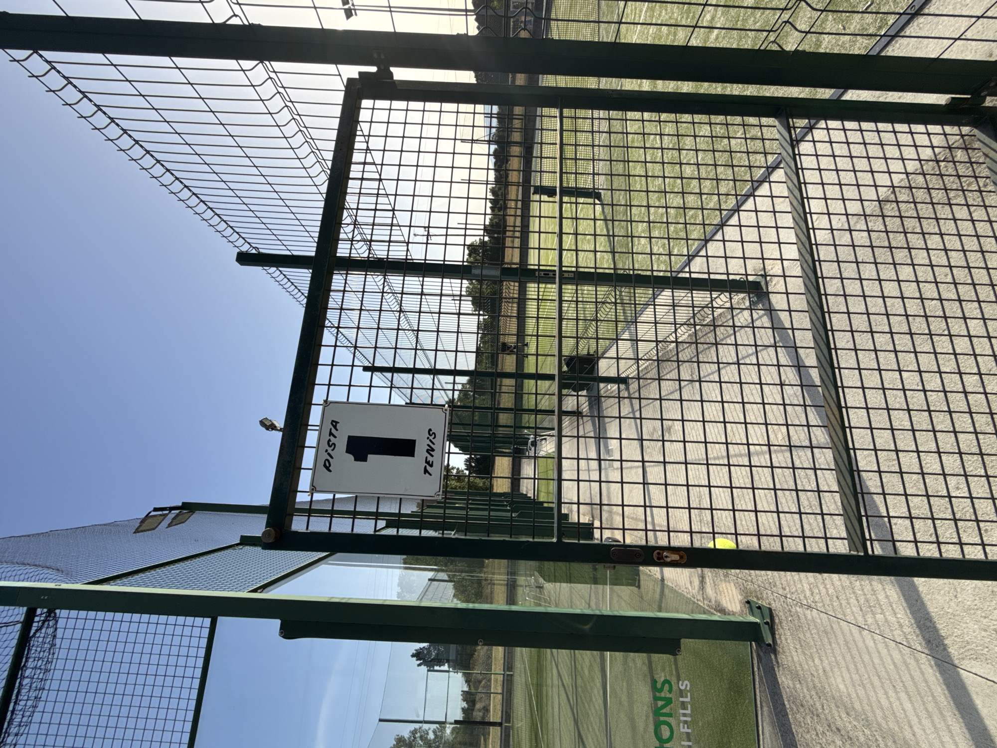 The image shows a fenced-off tennis court in Vidreres, Spain. A sign on the gate reads "PISTA 1 TENIS", indicating it is Court 1 for tennis. The court is made of green artificial turf, and a net is visible. The sky is clear and blue, with the sun setting in the background, casting long shadows. The scene appears to be a sports facility, possibly a public park or a private club, with other courts visible in the distance. There are no people in the immediate foreground of the image. The overall impression is one of a well-maintained outdoor sports venue on a sunny day.