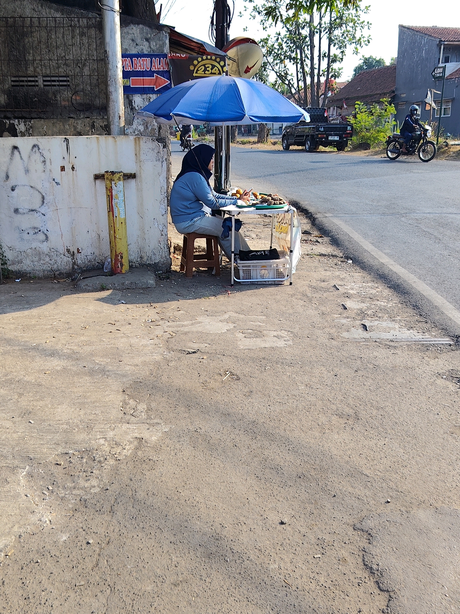 The image depicts a street scene in Purwakarta, Indonesia. A person wearing a blue hijab and a light blue long-sleeved shirt and pants is seated at a small, portable food stall under a blue umbrella. The stall has a white canopy and displays food items on a tray. A brown plastic stool is used by the vendor. The stall is situated on a dirt and asphalt roadside. In the background, a black pickup truck is parked on the road, and a person on a motorcycle is riding by. Buildings and trees are visible further in the background, suggesting a semi-urban or roadside environment. A sign with Indonesian text and an arrow pointing to the right is mounted on a wall behind the stall. The ground is littered with small debris. The lighting suggests it is daytime, with shadows cast by the objects.