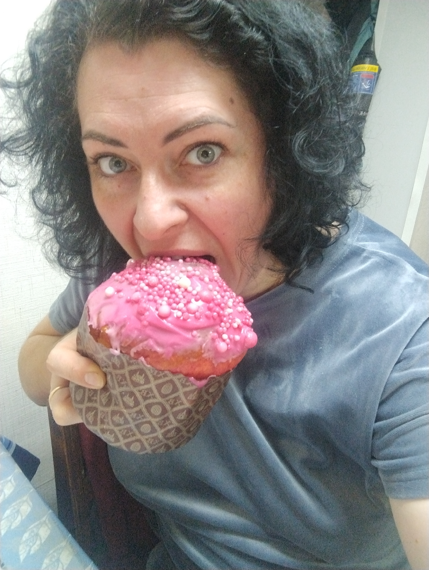 A woman with dark curly hair is holding a decorated cake to her mouth, apparently taking a large bite. The cake has pink icing and is covered in small, light-colored sprinkles. She is wearing a grayish-blue, short-sleeved velvet shirt. The location is assumed to be Grishanki, Russia, based on information not visible in the image.