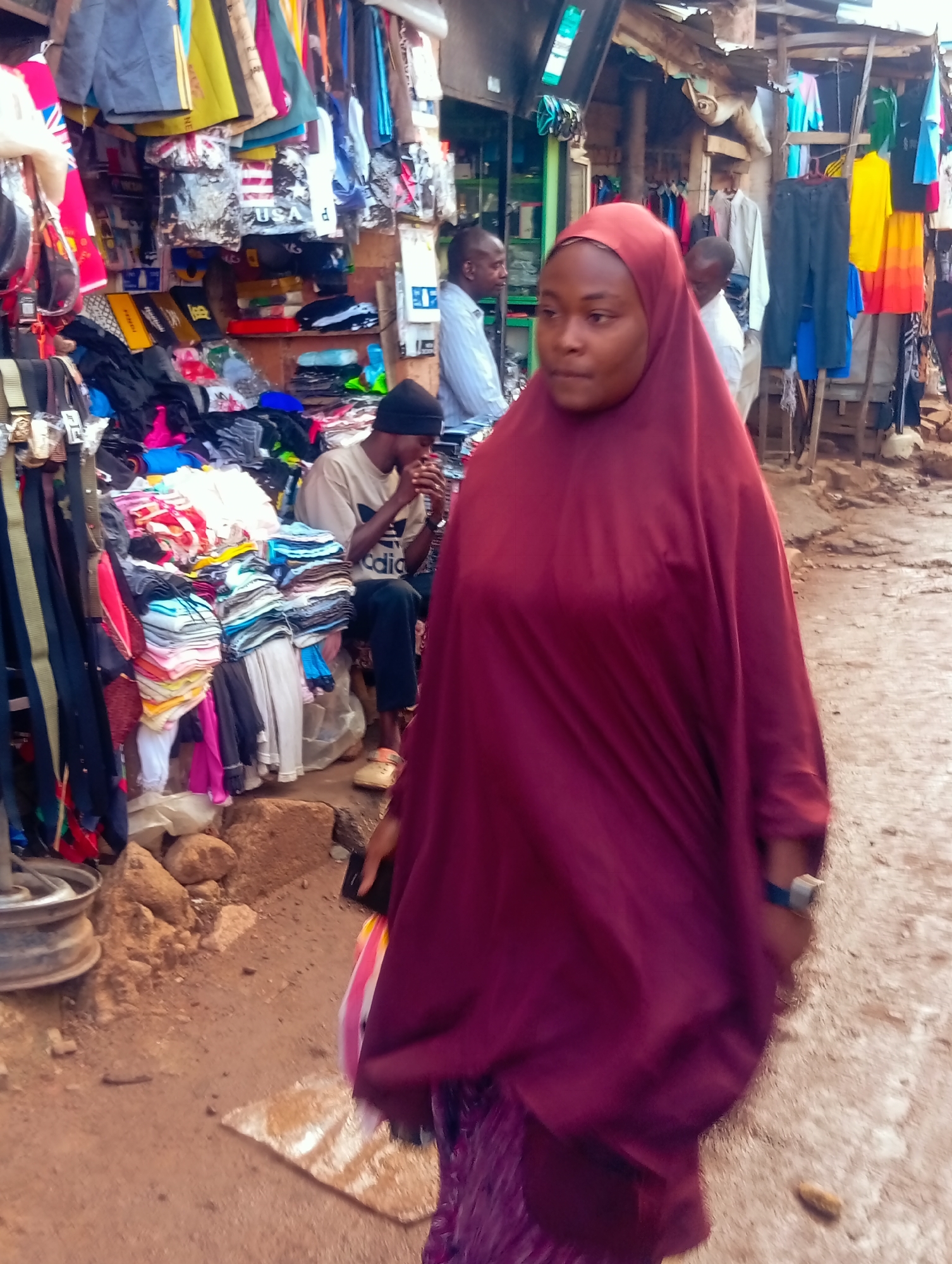 The image shows a street market scene in Bukuru, Nigeria. The primary subject is a woman wearing a dark red hijab and a matching dress, walking towards the viewer. She is carrying a shopping bag. Behind her, other individuals are visible, including a man in a light-colored shirt and a black beanie, who appears to be seated at a stall. Several stalls are packed with merchandise, primarily clothing and accessories. Items include folded stacks of shirts, pants, belts, and what appear to be t-shirts with various graphic prints, some with text like "USA". The stalls have a makeshift construction, with corrugated metal roofing and wooden structures. The ground is dirt and gravel, suggesting an outdoor market. The lighting suggests it is daytime, with the sun casting shadows. The overall atmosphere is that of a busy, everyday market environment.