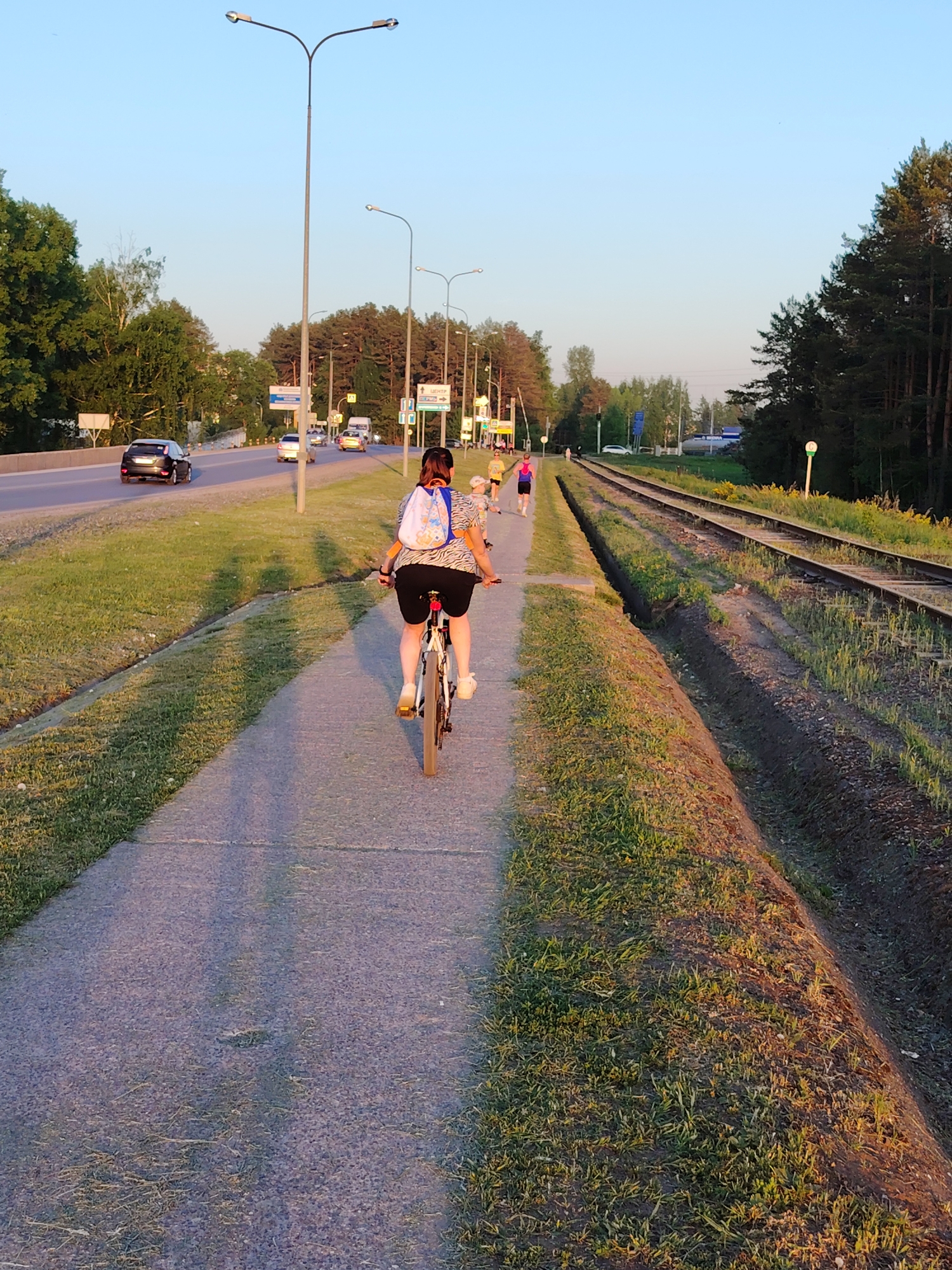 A cyclist rides on a paved path adjacent to railroad tracks in Chaikovsky, Russia, at sunset. The path runs parallel to a multi-lane highway with cars traveling on it. Several pedestrians are further ahead on the path. Grass grows on either side of the path and along the railroad tracks. Streetlights are visible along the highway. Trees border the road and the right side of the railroad tracks.