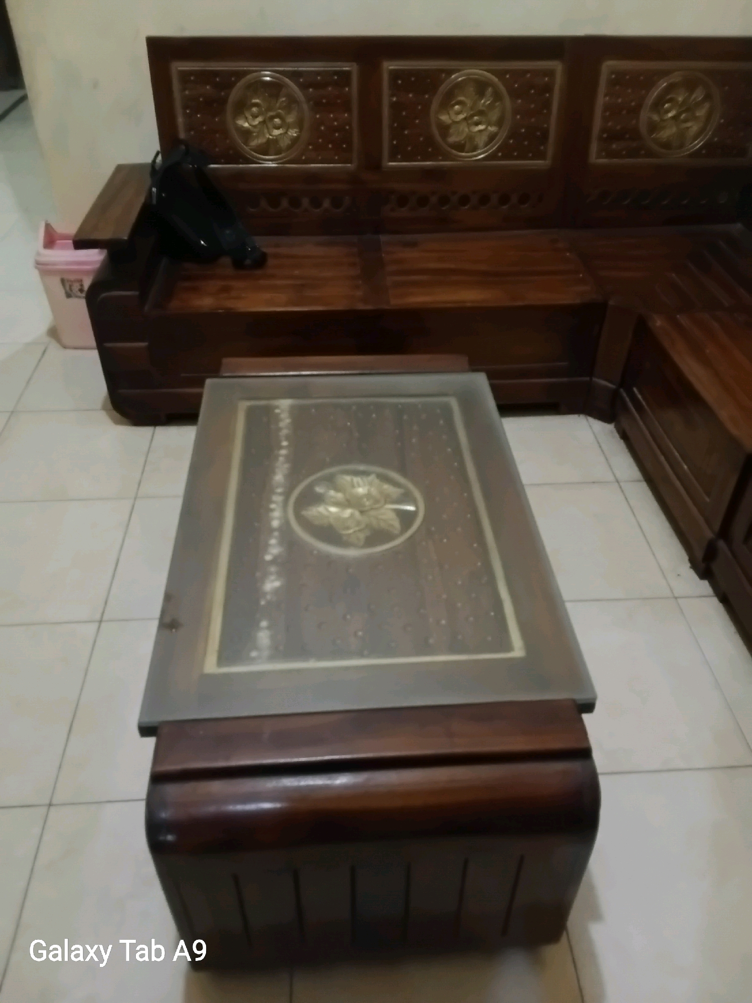 A low-angle, medium shot captures a dark wood corner sofa with a matching coffee table in what appears to be a residential indoor setting in Trenggalek, Indonesia. The sofa features decorative golden circular emblems with floral motifs on its backrest and has a dark wood finish. A black object, possibly a handbag or a shoe, rests on the left side of the sofa's seating area. To the left of the sofa, a light pink trash bin is partially visible. The coffee table, positioned in front of the sofa, has a glass top with a decorative golden floral emblem in the center. The table's base is constructed of dark wood with vertical slats. The floor is tiled in a light color with a grid pattern. The overall lighting is dim, suggesting it might be evening or an indoor space with limited natural light. The text "Galaxy Tab A9" is overlaid in the bottom left corner of the image.