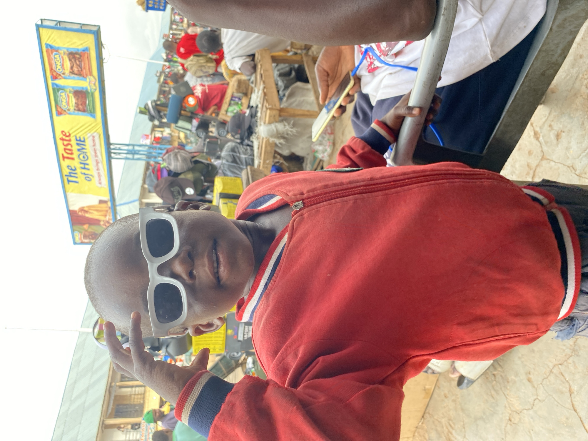The image depicts a child wearing white sunglasses and a red jacket, posing for a photo. The picture appears to be taken from a low vantage point. In the background, there is a crowded street scene with people, stalls, and merchandise. A sign advertising "The Taste of Home" is visible in the upper left. The location is Bukuru, Nigeria. Some people are seated on a bench and looking at a phone.