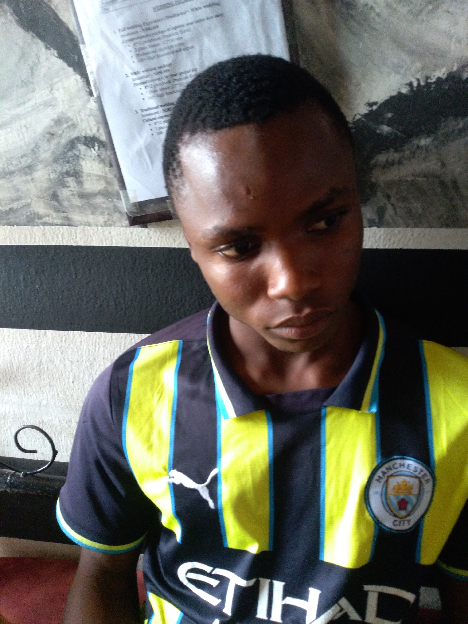 A close-up, head-and-shoulders shot depicts a young Black male. He is looking downwards and to the right, with his expression appearing thoughtful or somber. His dark, short hair is neatly cut. He is wearing a dark blue polo shirt with bright yellow and thin blue stripes running vertically down the front. The shirt features a Puma logo on the left chest and the Manchester City Football Club crest on the right. A sheet of paper with text is visible behind and to the left of his head, appearing to list wedding packages. The background consists of a striped pattern of black and white textured surfaces. A decorative black metal element with a spiral shape is visible on the left side of the frame.