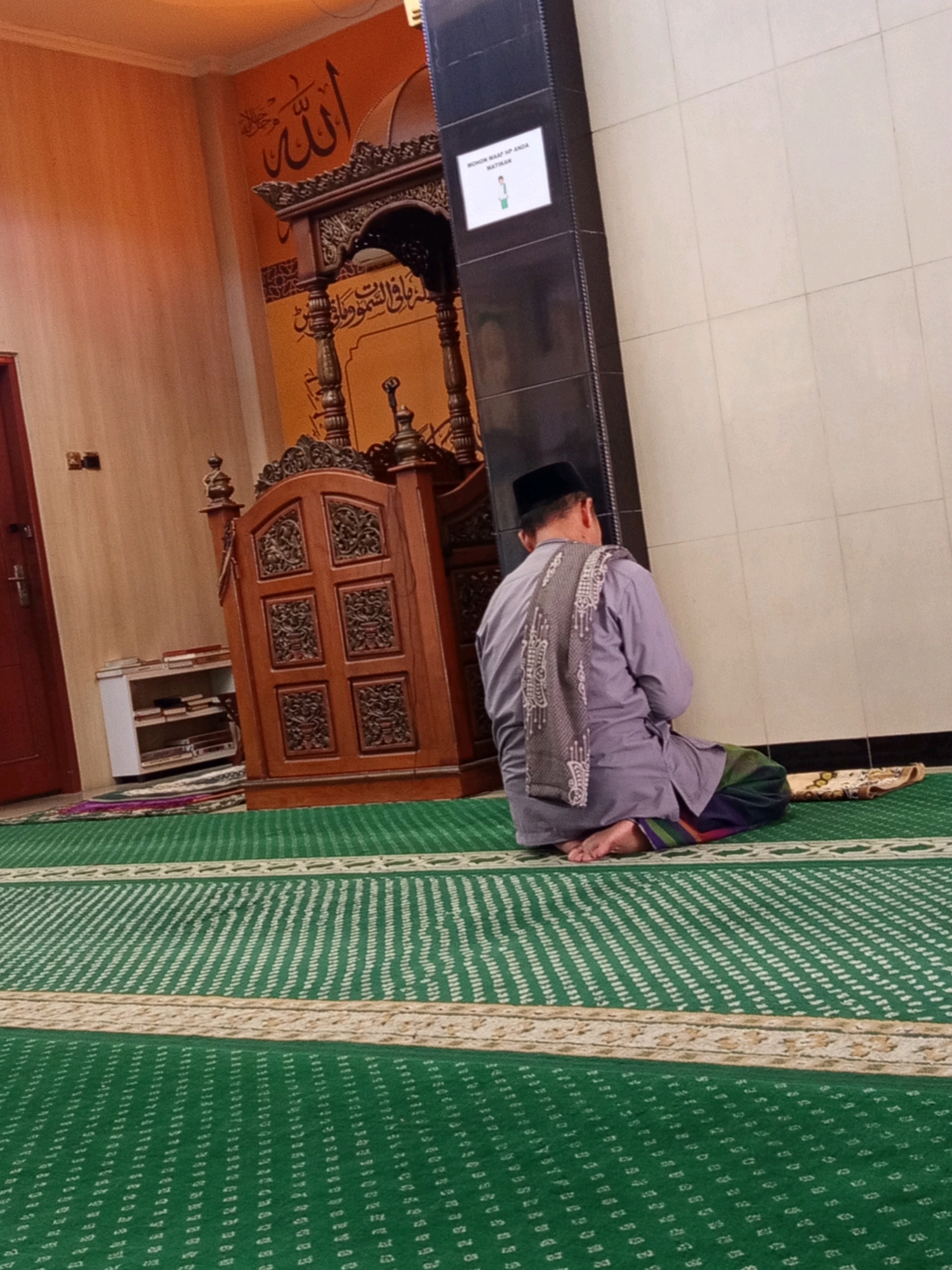 A man is sitting on the floor of a mosque, facing away from the camera. He is wearing a grey shirt, a black cap, and a sarong. He is looking towards a minbar, a raised platform used for sermons in a mosque. The mosque has green carpeted floors with white geometric patterns. There is a wooden minbar with intricate carvings. In the background, there is an orange wall with Arabic calligraphy. A small white shelf with books is visible to the left of the minbar. A black pillar with a white sign is next to the man. The image appears to be taken in a mosque in Karawang, Indonesia, given the context provided about the location.