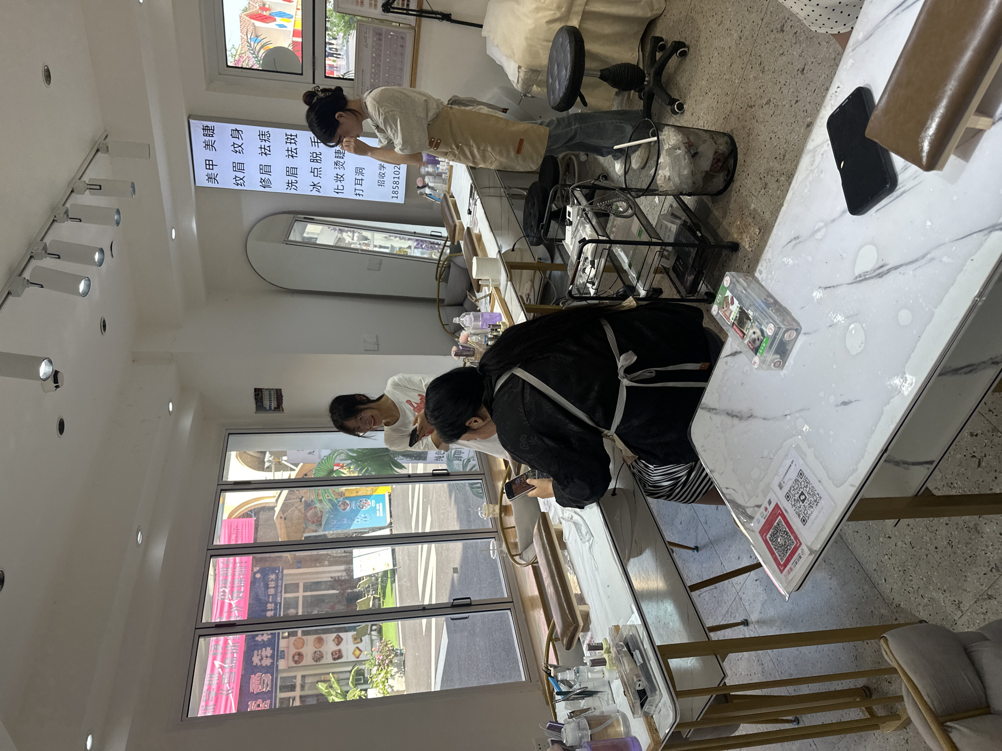Here is a description of the image in detail:
**Location:** Chongqing High-tech Zone, China
**Scene:** The interior of a beauty salon, possibly specializing in nail care and eyebrow services. Two women are present. One woman is standing at a counter and appearing to be working on something in front of her. The second woman is standing nearby and is also working at a counter. There are two manicure stations.
**Subjects:** Two women, presumably salon staff, are engaged in some type of work. One woman is wearing a beige apron and the second woman is wearing a black shirt and an apron.
**Objects:** There are manicure tables. There are various bottles and tools on the tables, typical of a beauty salon. A menu can be seen advertising various services. There is a trash can present. There is a set of large windows looking out onto the street.
**Actions/Interactions:** The women are working on tasks associated with the beauty salon. They both are working at the tables near beauty products and tools.
**Environment:** The interior is well-lit with modern lighting fixtures. The decor is clean and minimalist. Through the window, the street scene of Chongqing High-tech Zone is visible.